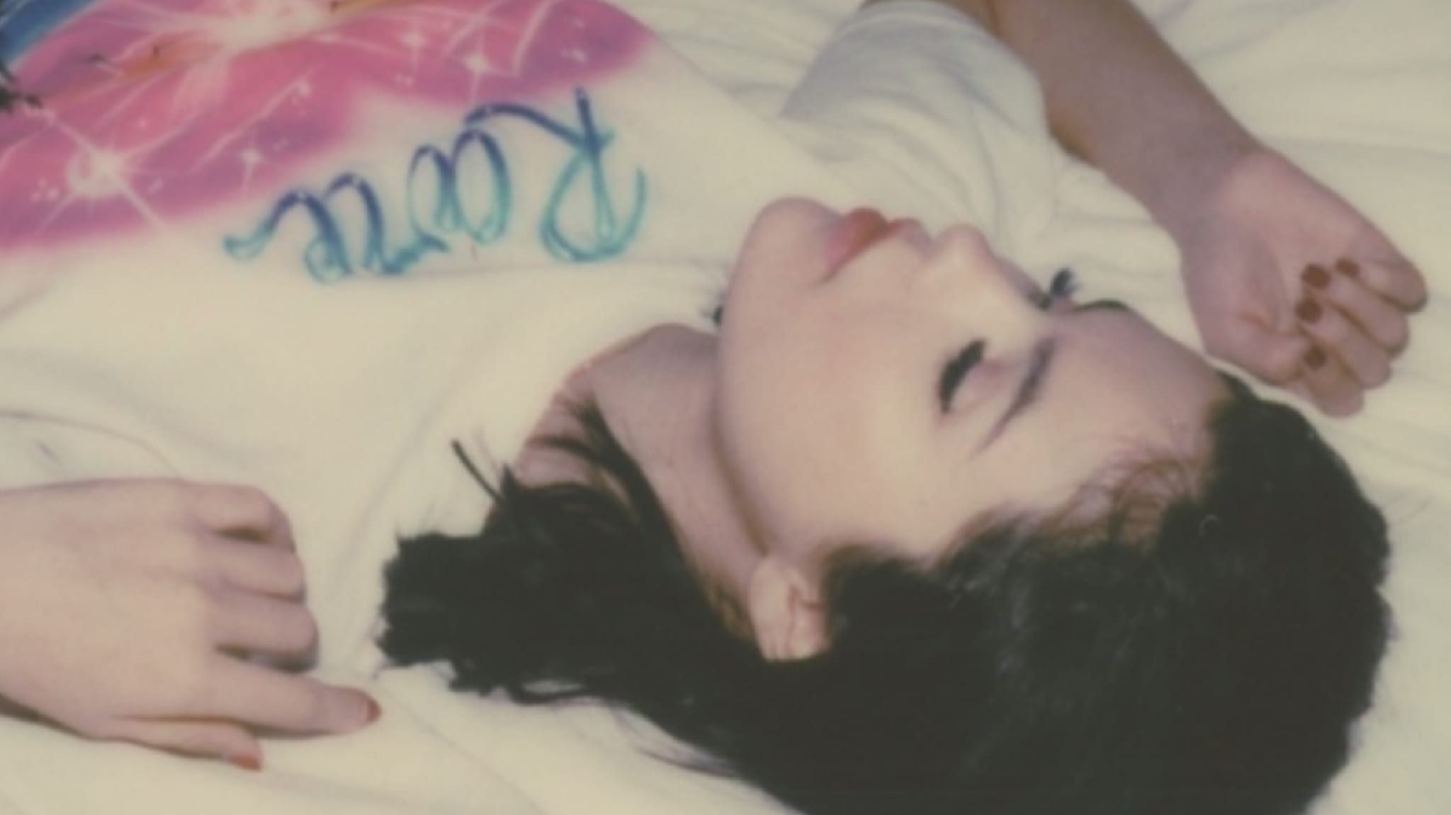 Selena Gomez lies on a bed, wearing a white shirt with colorful text. The image has a soft, dreamy quality.