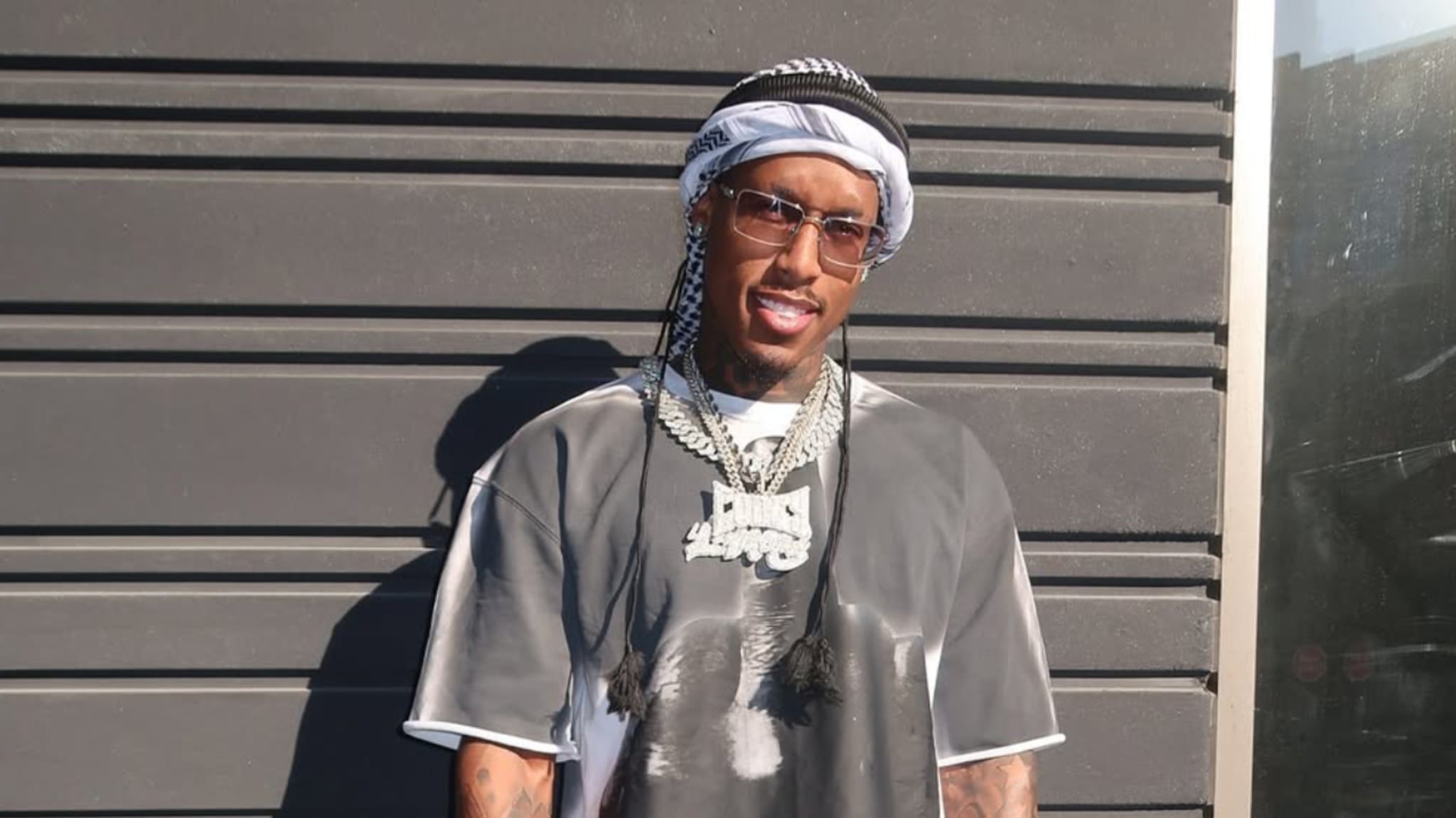 A man wearing a headscarf, sunglasses, and a large chain necklace, standing in front of a dark wall.