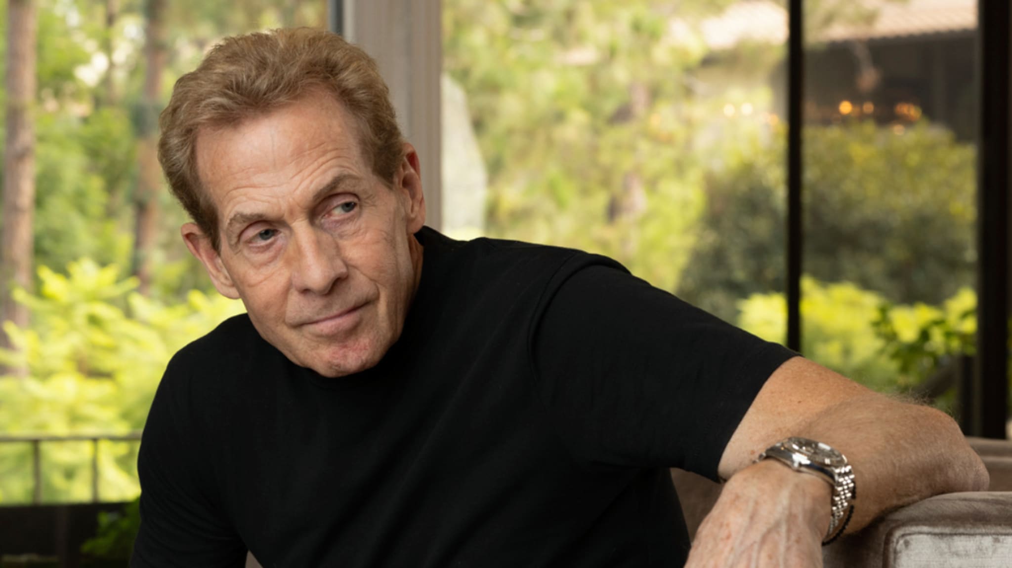 Skip Bayless with short light brown hair wearing a black shirt, sitting indoors with a window view of greenery in the background.