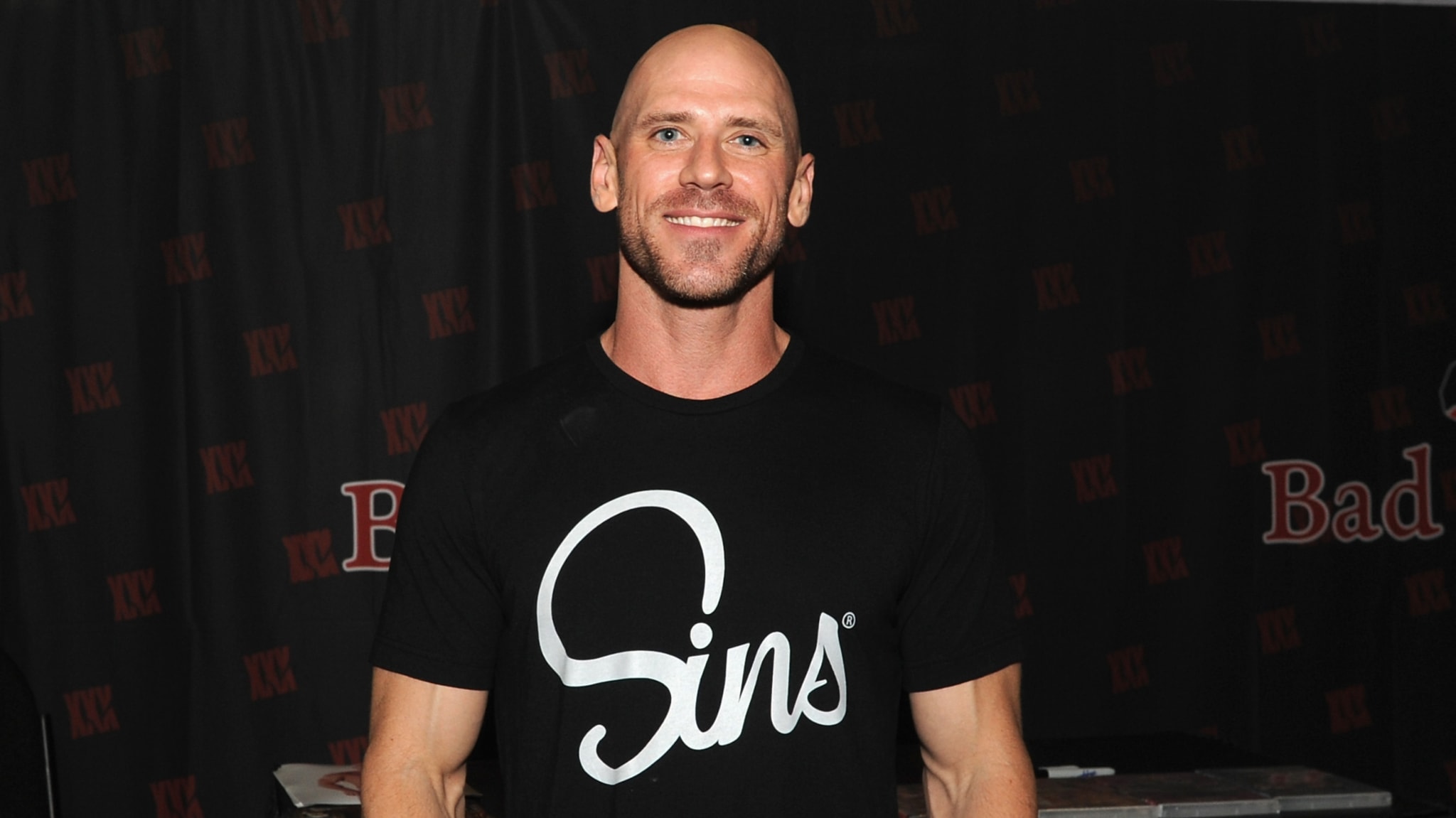A man with a shaved head and beard wearing a black T-shirt with "Sins" written on it, standing in front of a dark background.