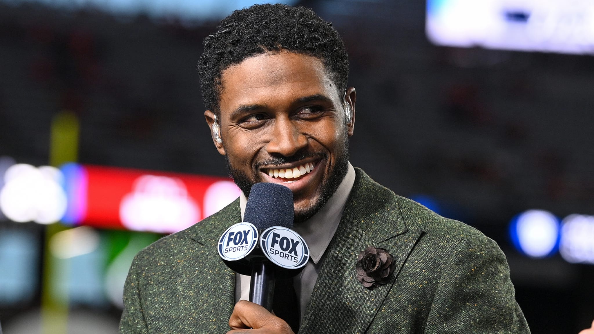 Reggie Bush in a suit smiles while holding a Fox Sports microphone.