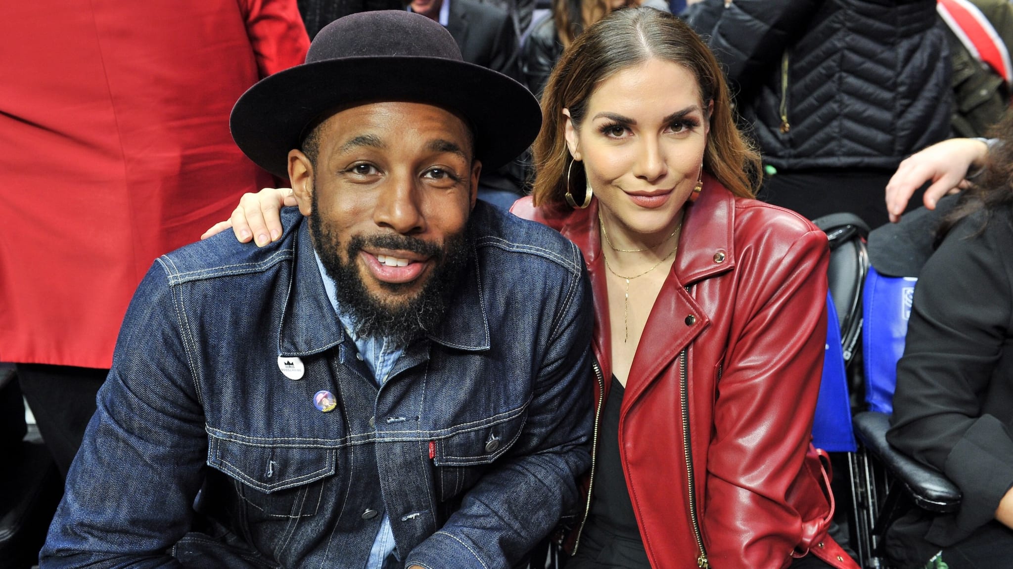 A man and woman sitting together, both smiling. The man wears a denim jacket and hat, and the woman wears a red leather jacket.
