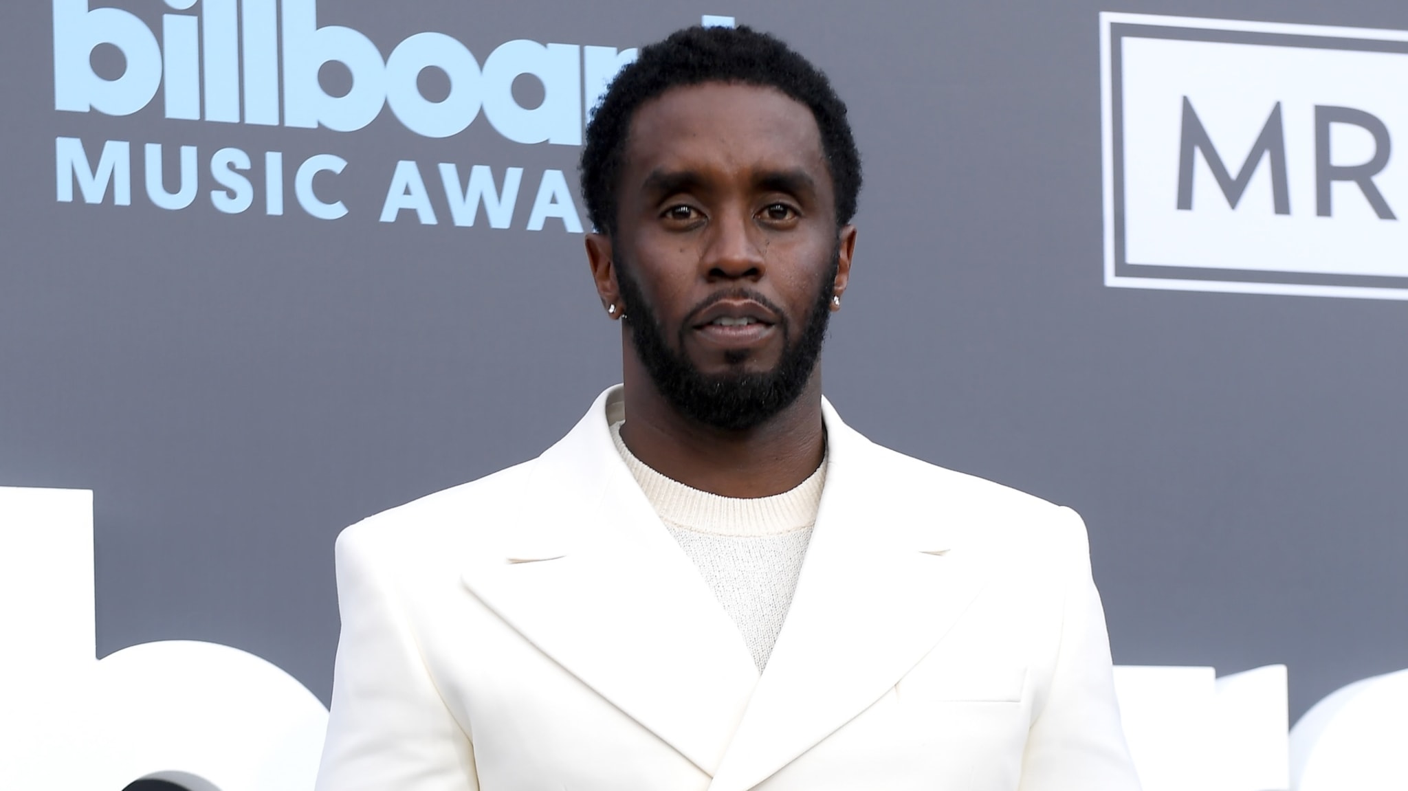Sean "Diddy" Combs at the Billboard Music Awards, wearing a white suit, standing against the event's backdrop.
