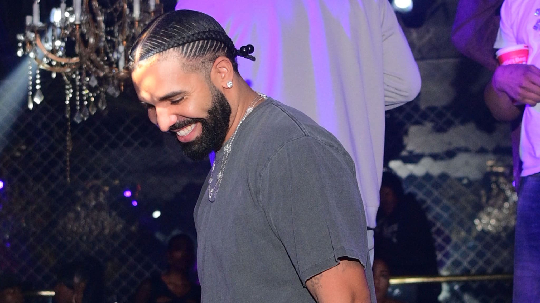 Drake smiling at a party, wearing a dark shirt with braided hair. Chandelier and people in the background.