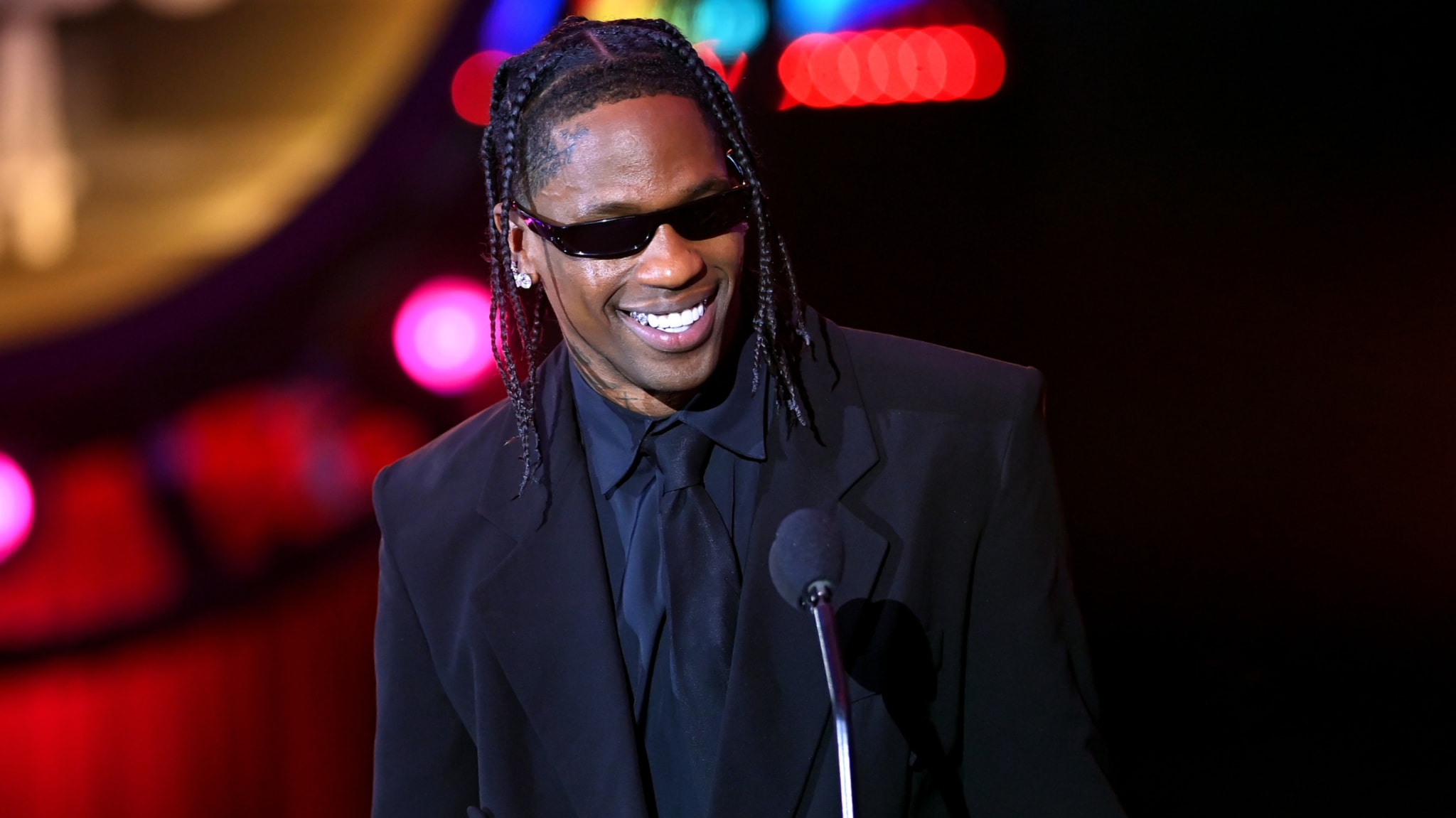 Travis Scott wearing sunglasses and a black suit, smiling at a microphone on stage with colorful lights in the background.