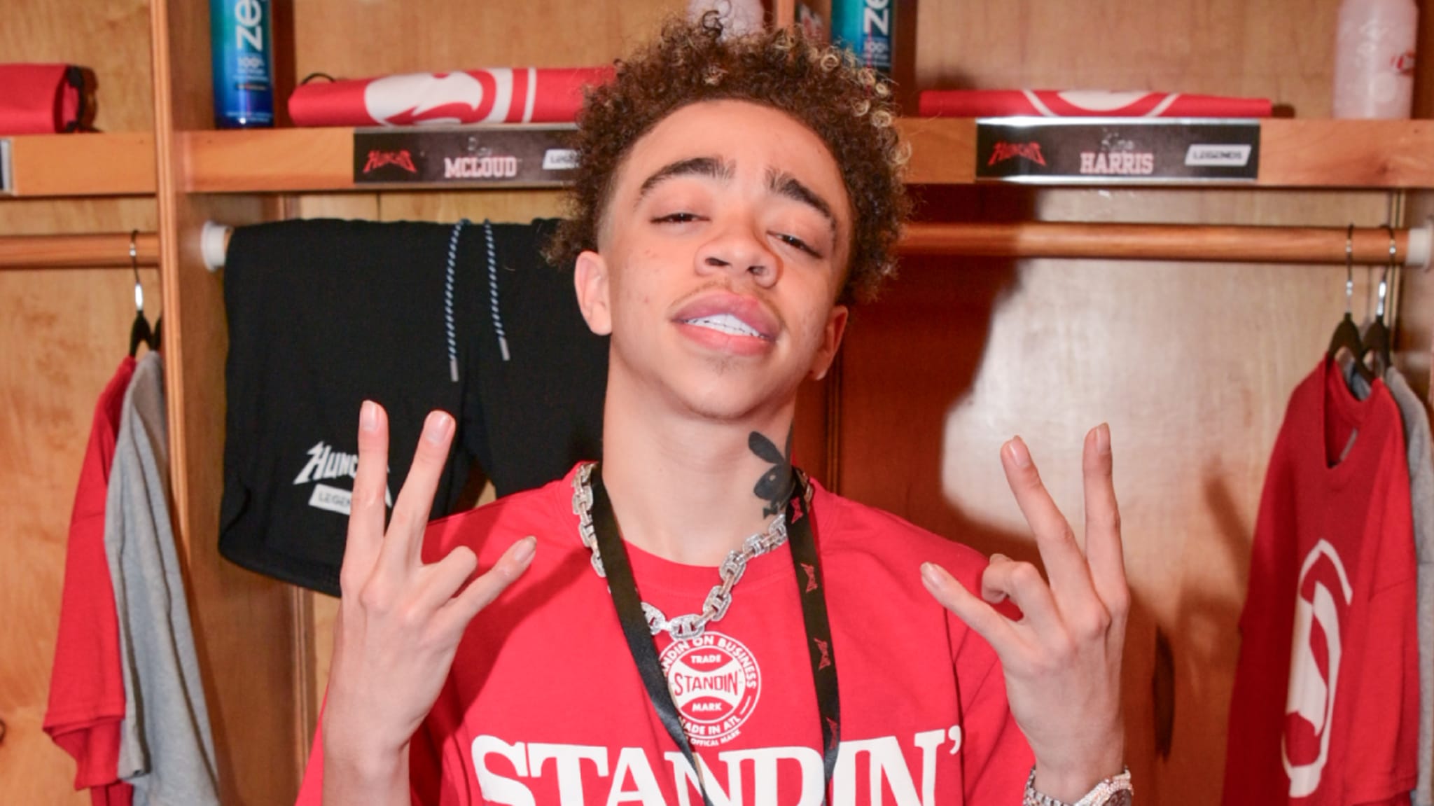 King Harris in a red shirt poses with hand gestures in front of a locker with clothing and towels.