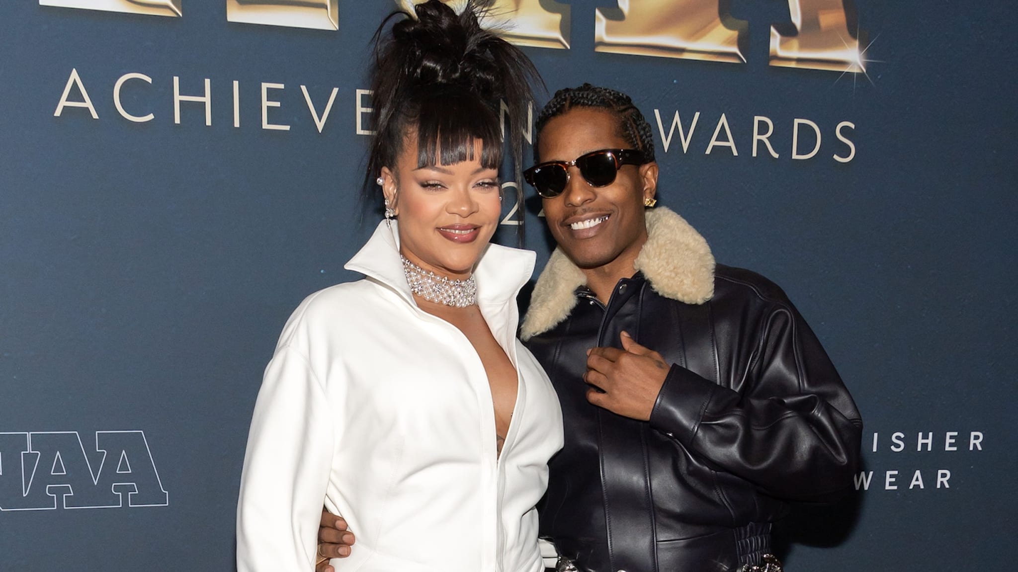 Rihanna and A$AP Rocky at an event, with Rihanna in a white outfit and A$AP Rocky in a dark jacket and sunglasses.