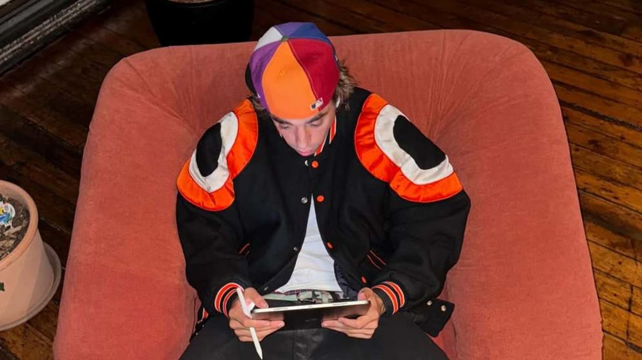 A person sits on a pink chair, wearing a colorful hat and black jacket with orange and white accents, using a tablet with a stylus.