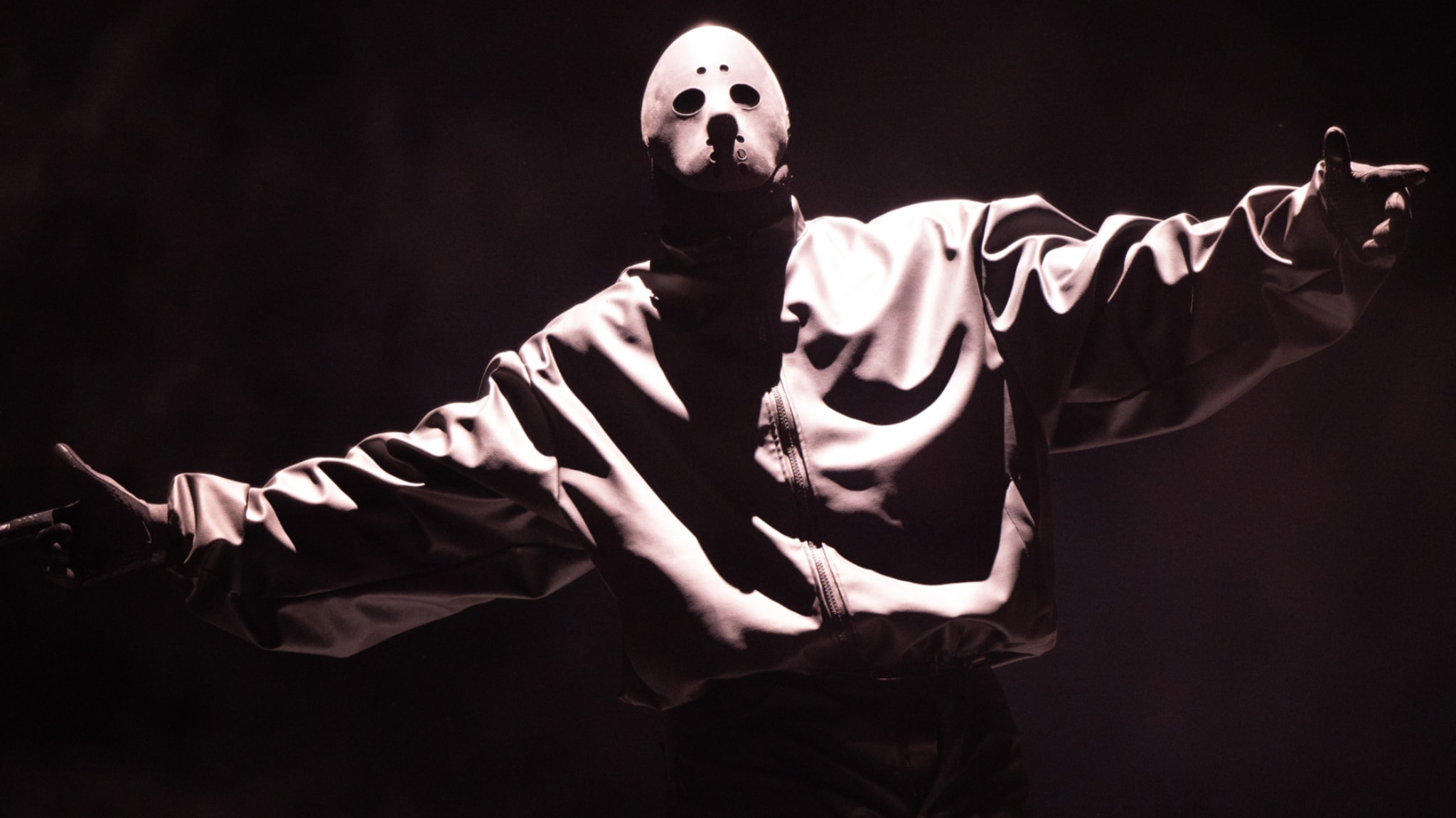 Ye wearing a mask with holes for eyes and mouth, dressed in a loose jacket, stands with arms outstretched in dramatic lighting.