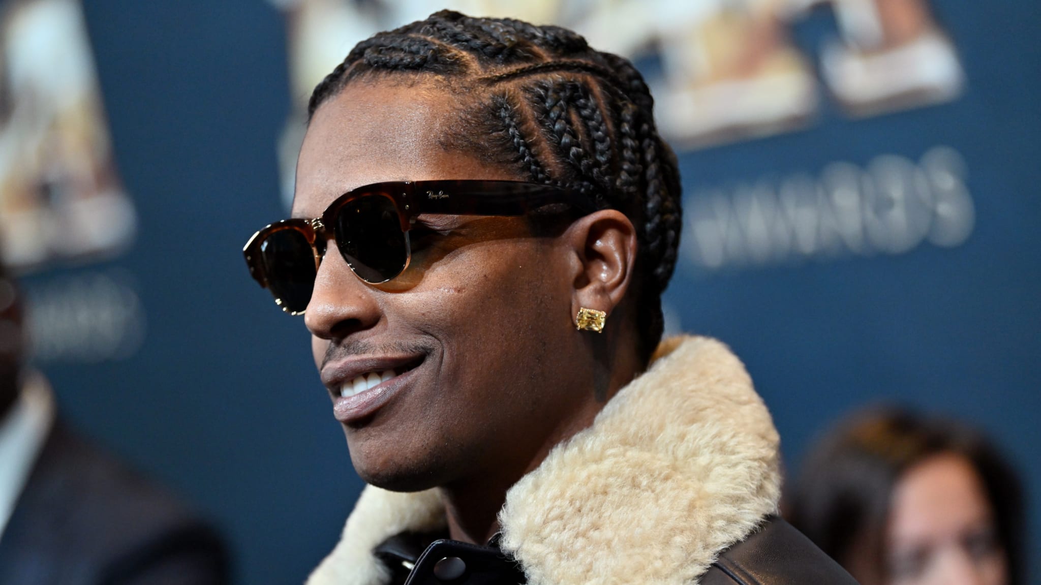 A$AP Rocky wearing sunglasses and a fur-collared jacket, smiling at an event.
