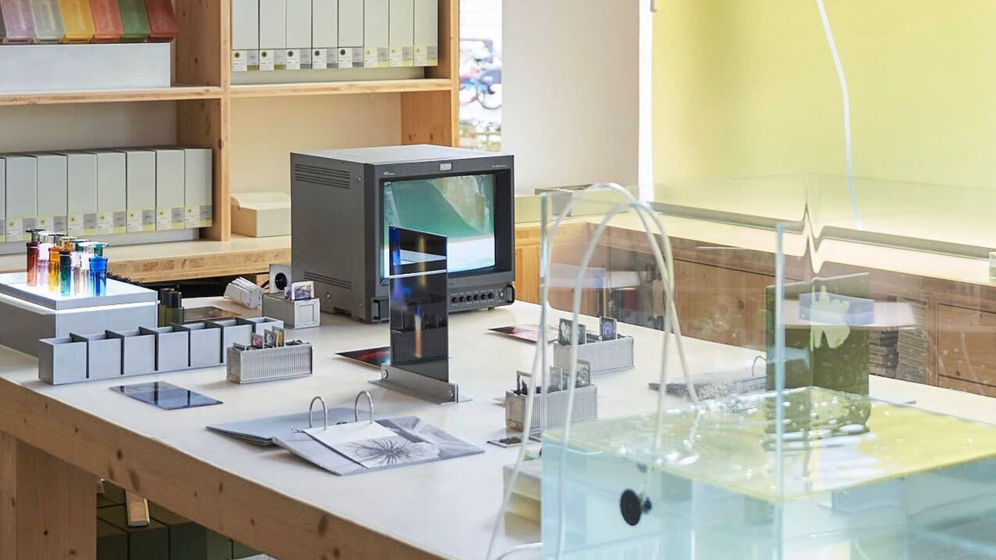 The Broken Arm Paris, A modern laboratory workspace with a monitor, test tubes, binders, and papers on a wooden table. Shelves with files are in the background.
