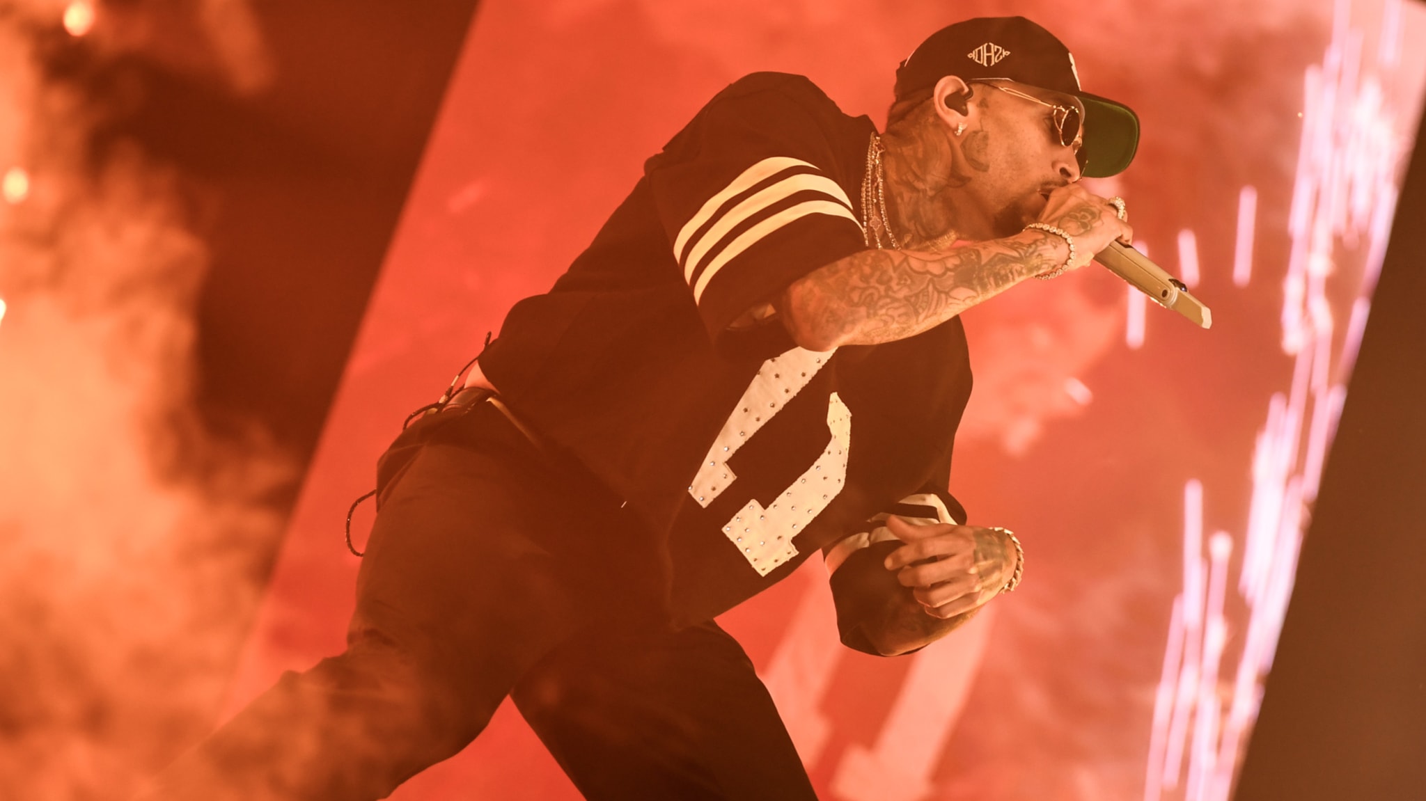 Chris Brown on stage with tattoos, wearing a black cap and jersey, holding a microphone. The background is illuminated with red lighting.