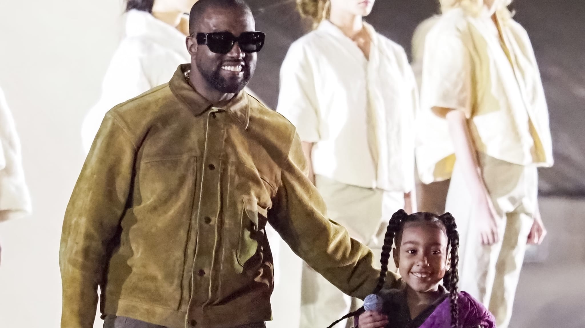 Kanye West wearing sunglasses and a brown jacket, smiling with North who's holding a microphone.