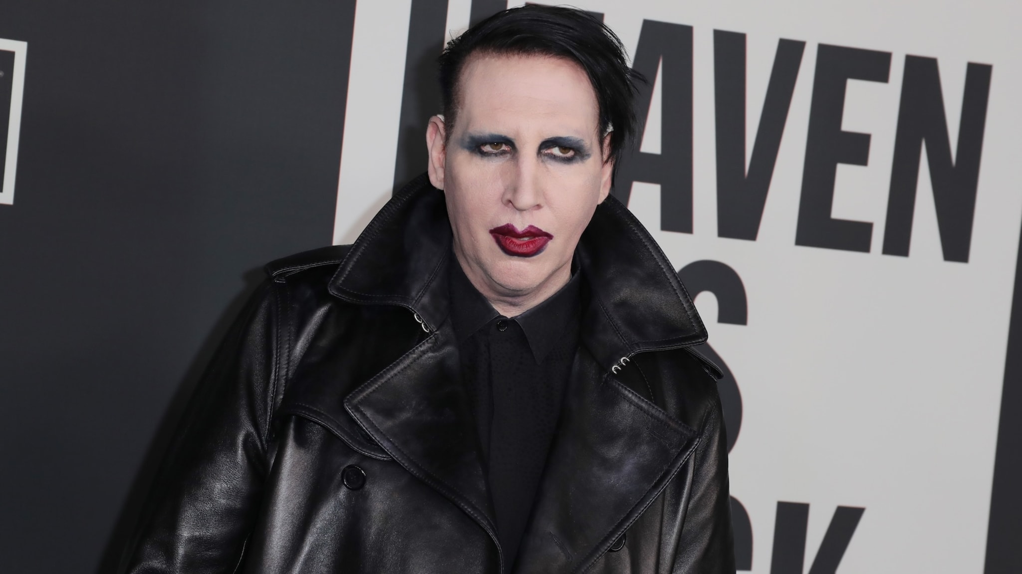 Marilyn Manson in a black leather coat, with dark makeup, at an event with a black and white background.
