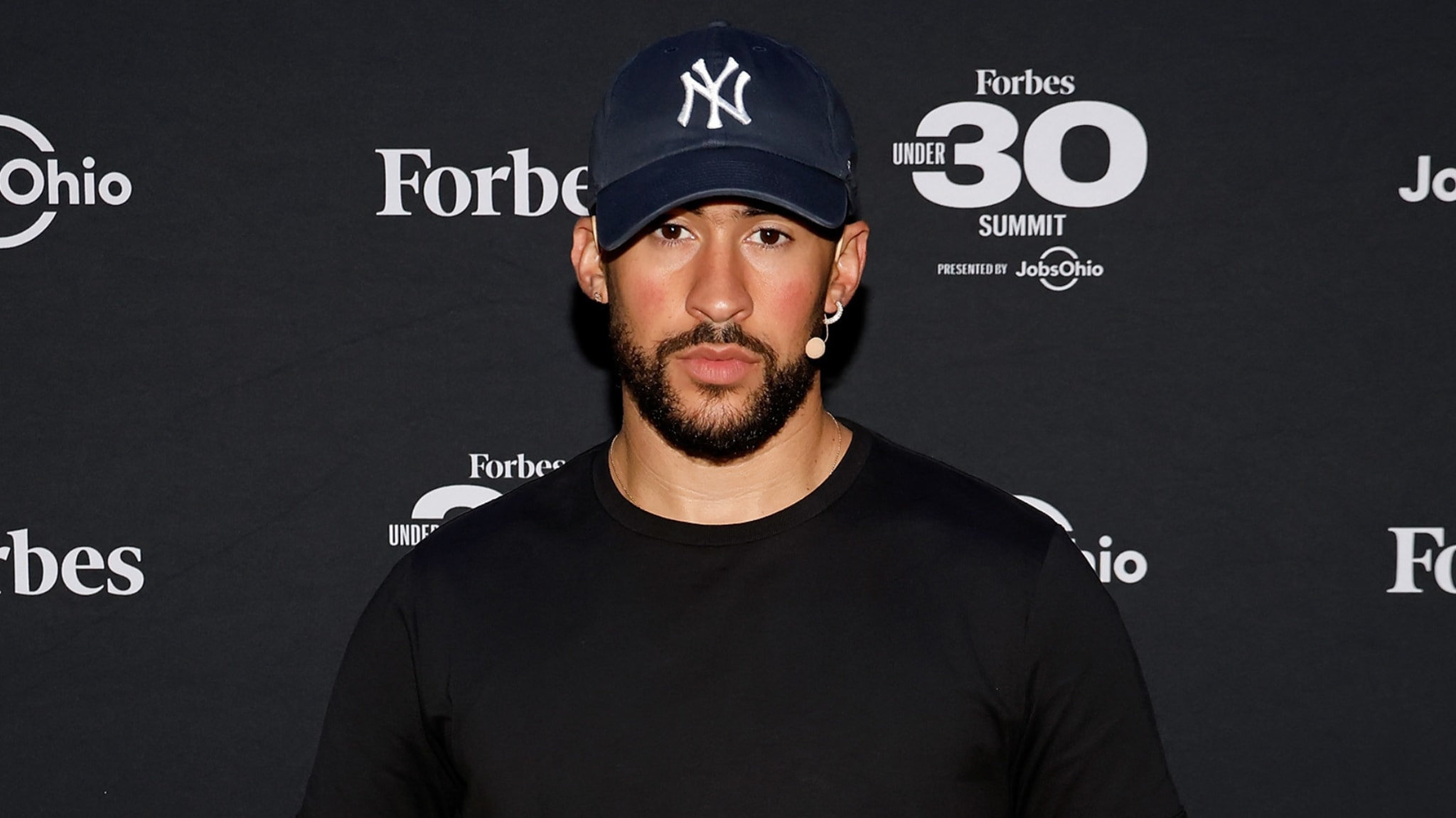 A man wearing a New York Yankees cap and a black shirt at the Forbes Under 30 Summit backdrop.