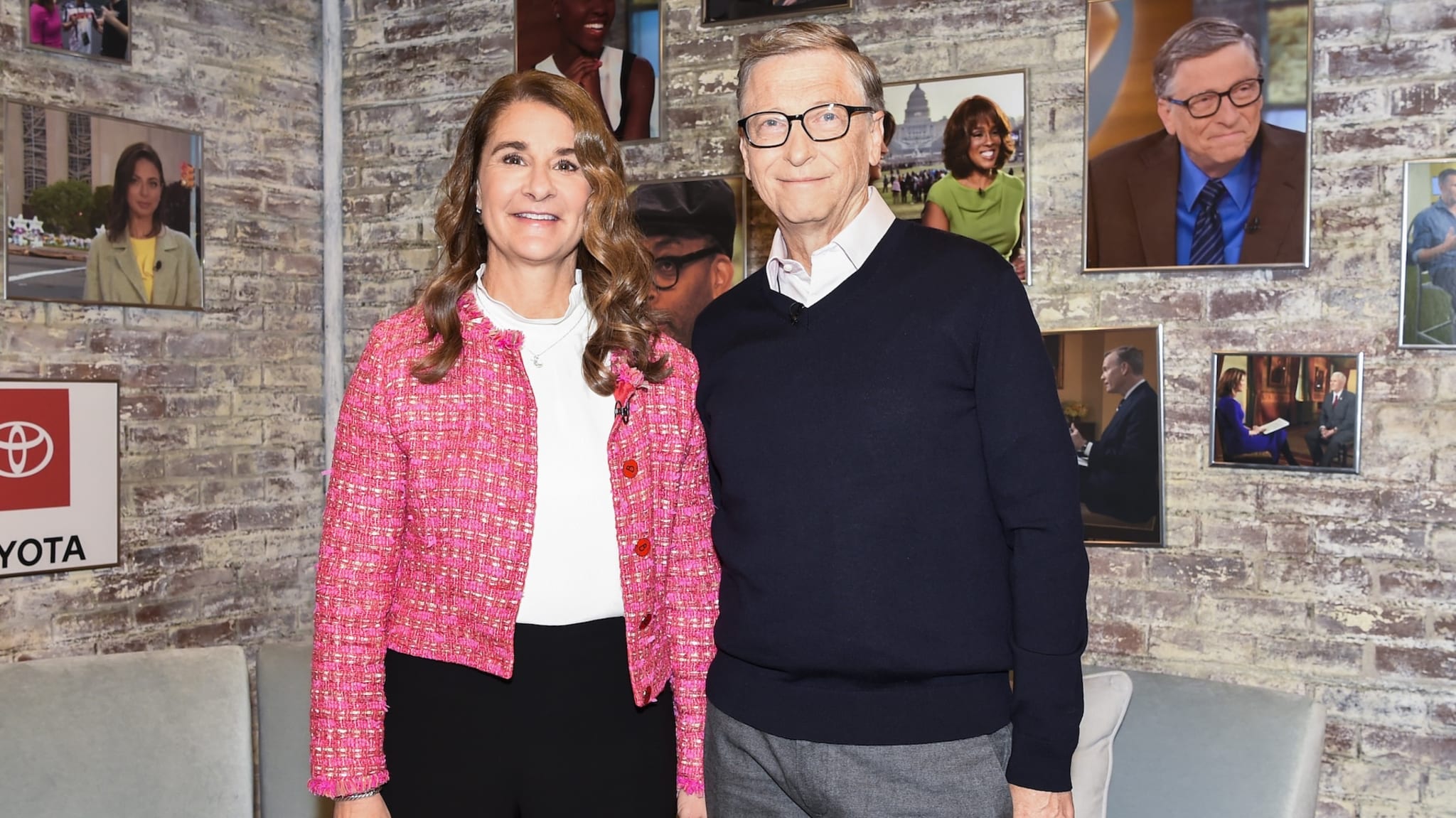 Bill Gates and Melinda Gates standing together, smiling, with a backdrop of framed photos on a brick wall.