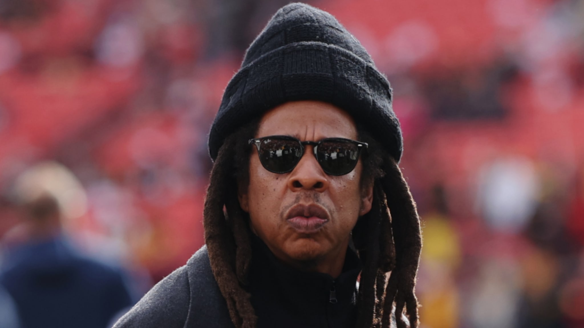 A person wearing sunglasses and a beanie, with long dreadlocks, standing outdoors with a blurry background.