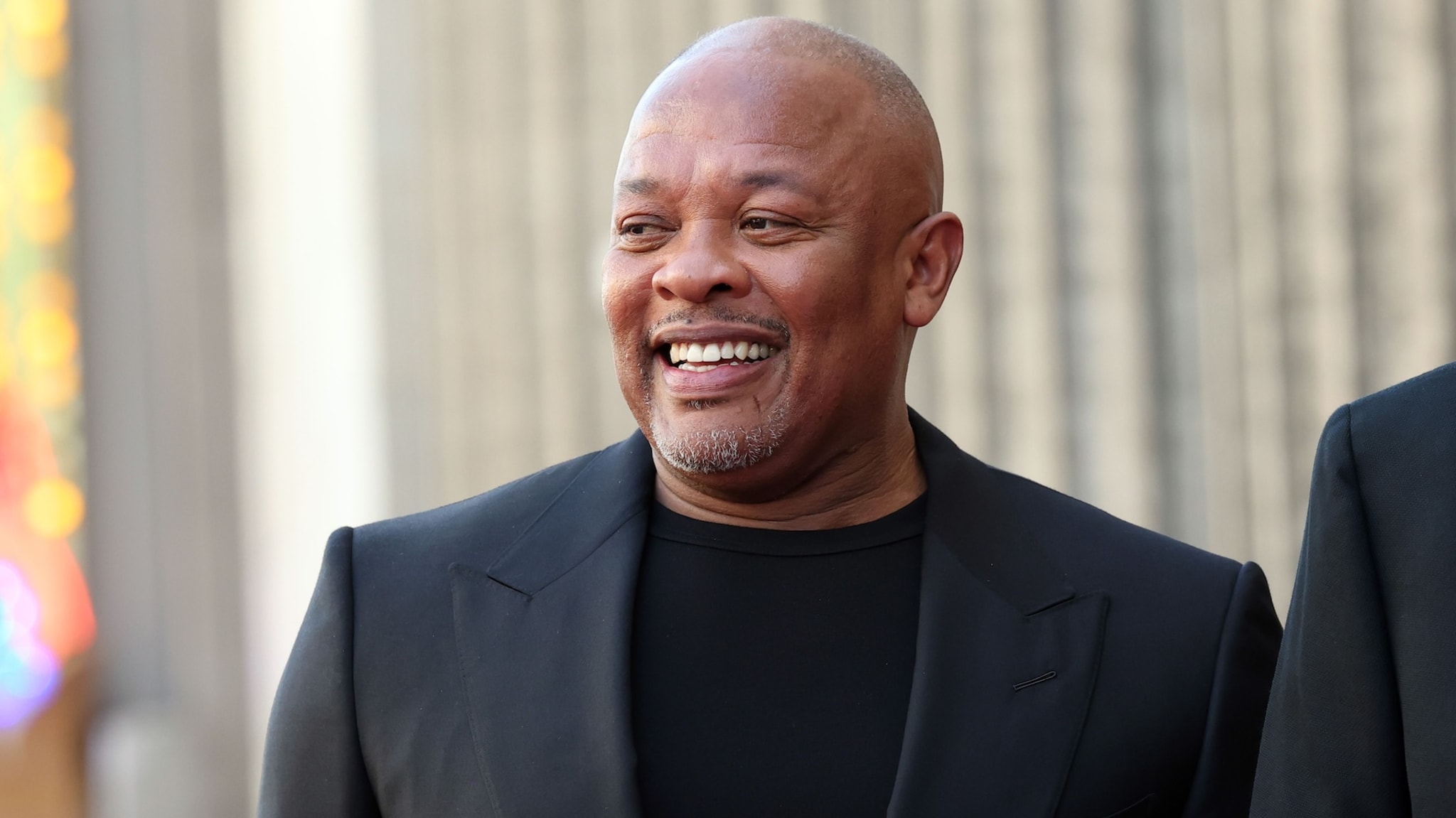 Dr. Dre in a black suit smiling, standing outdoors against a blurred background.