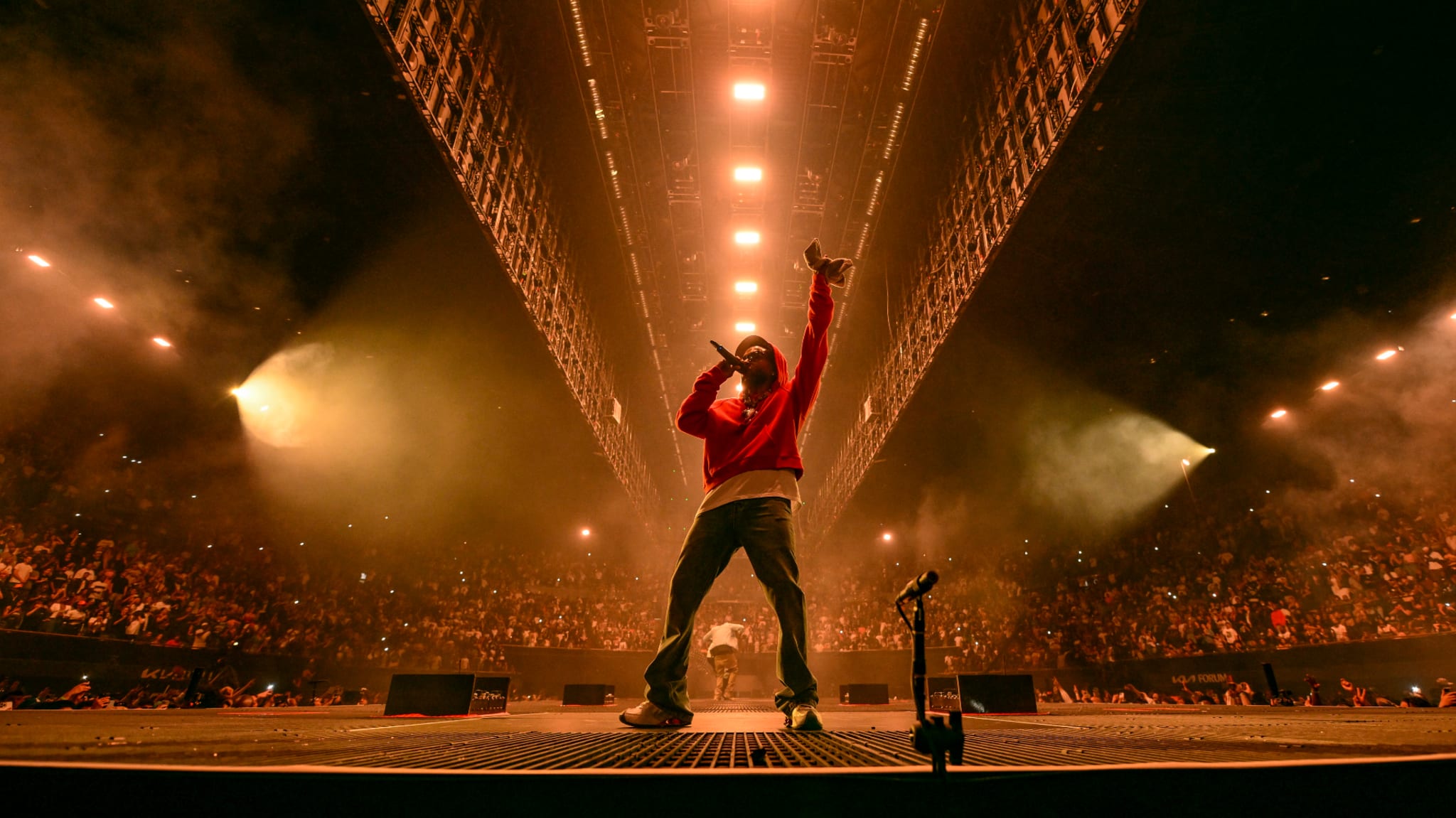 Kendrick Lamar on stage in a large, crowded arena, with dramatic lighting and smoke effects.