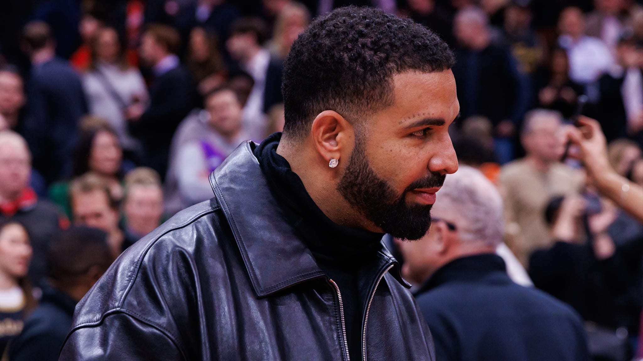 Drake wearing a black leather jacket, standing in a crowded area.