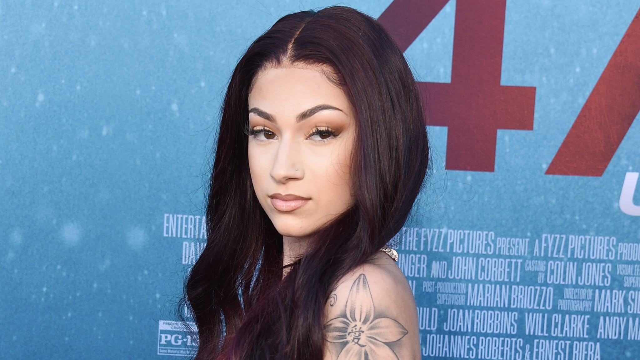 A young woman with long dark hair and a floral tattoo on her shoulder poses against a blue backdrop with text.