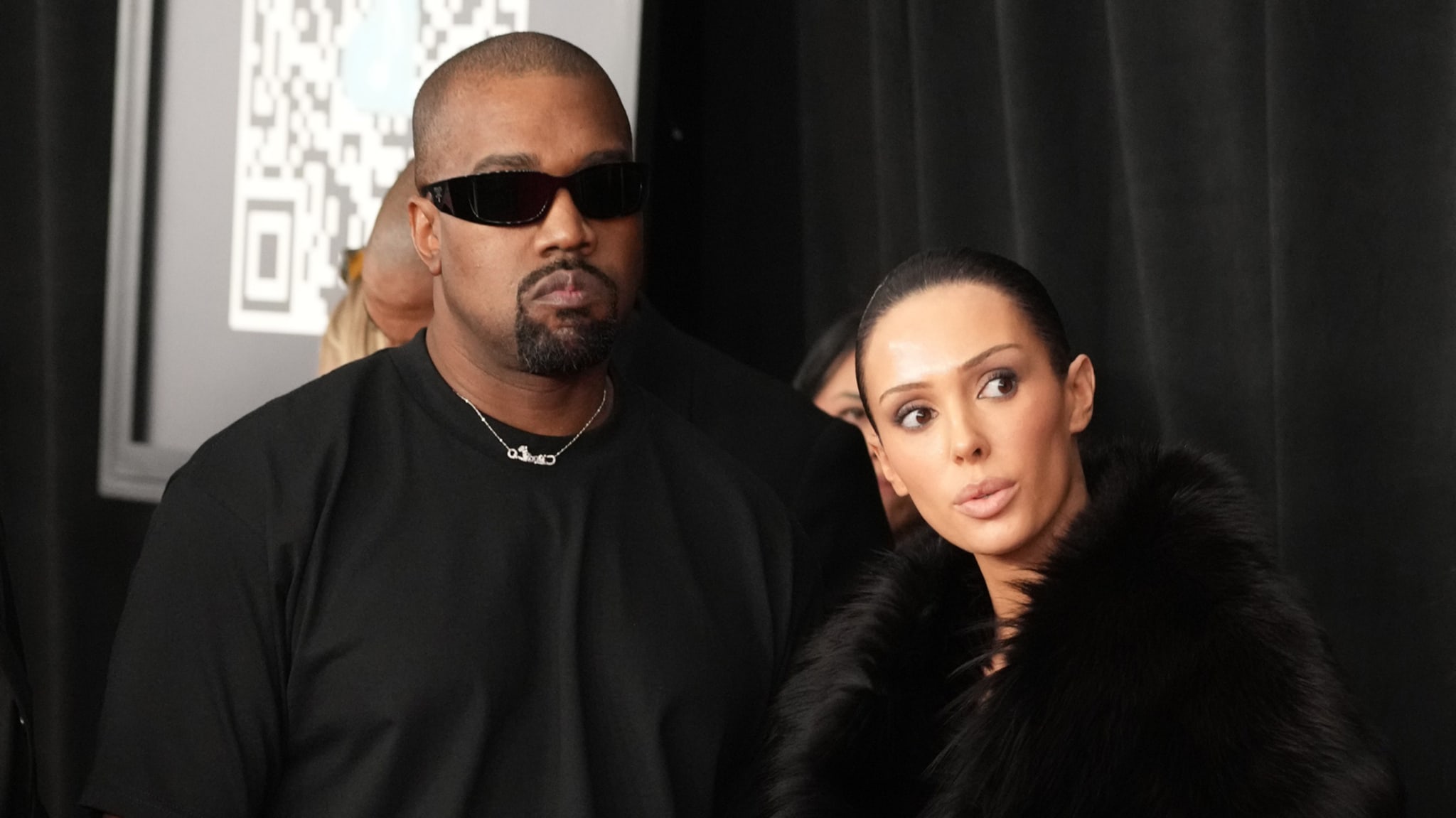 Kanye West wearing sunglasses, alongside Bianca in a fur coat, standing indoors.