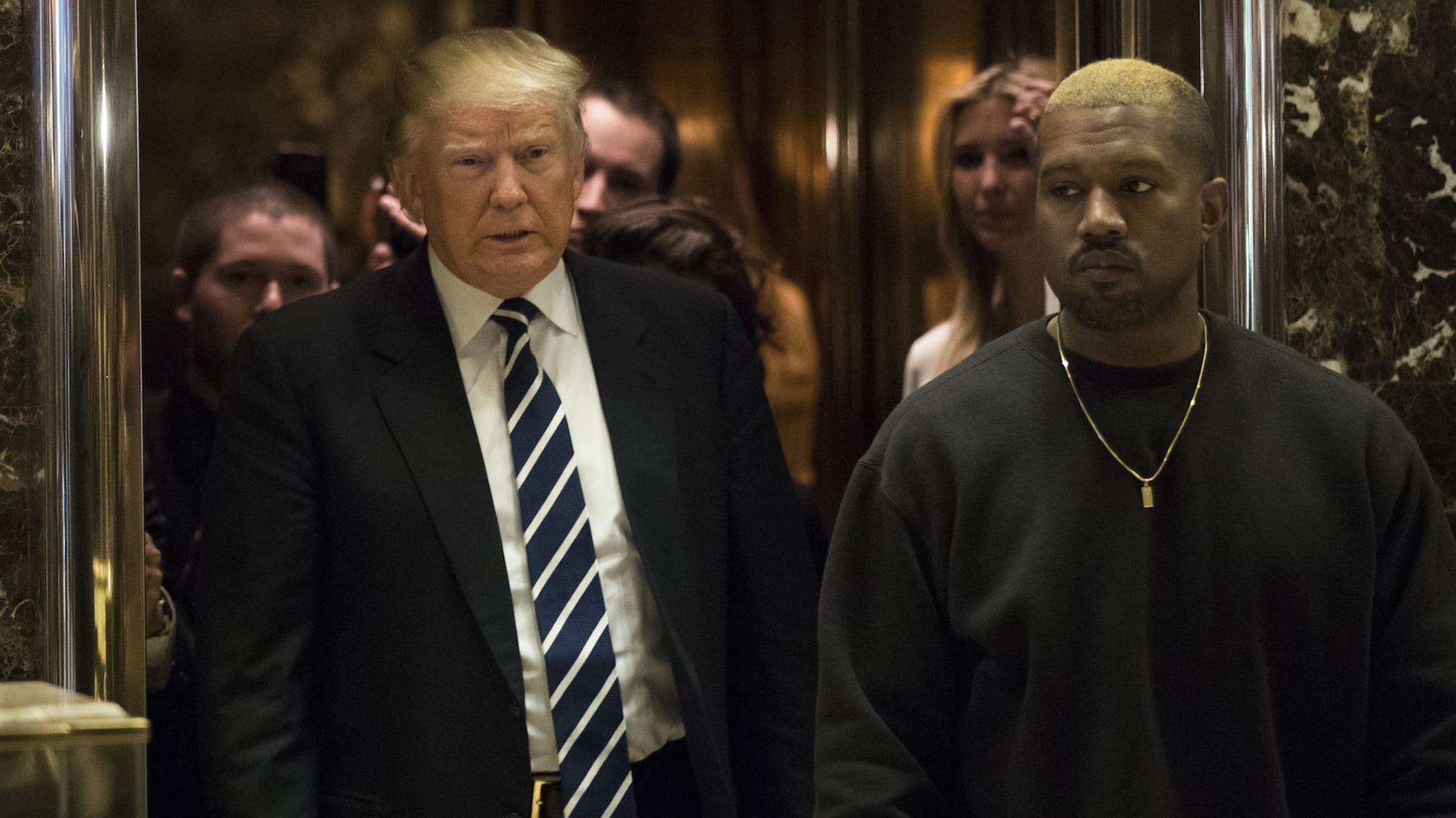 Donald Trump and Kanye West walking together, surrounded by people. Trump is in a suit and tie, Kanye in a dark sweatshirt.