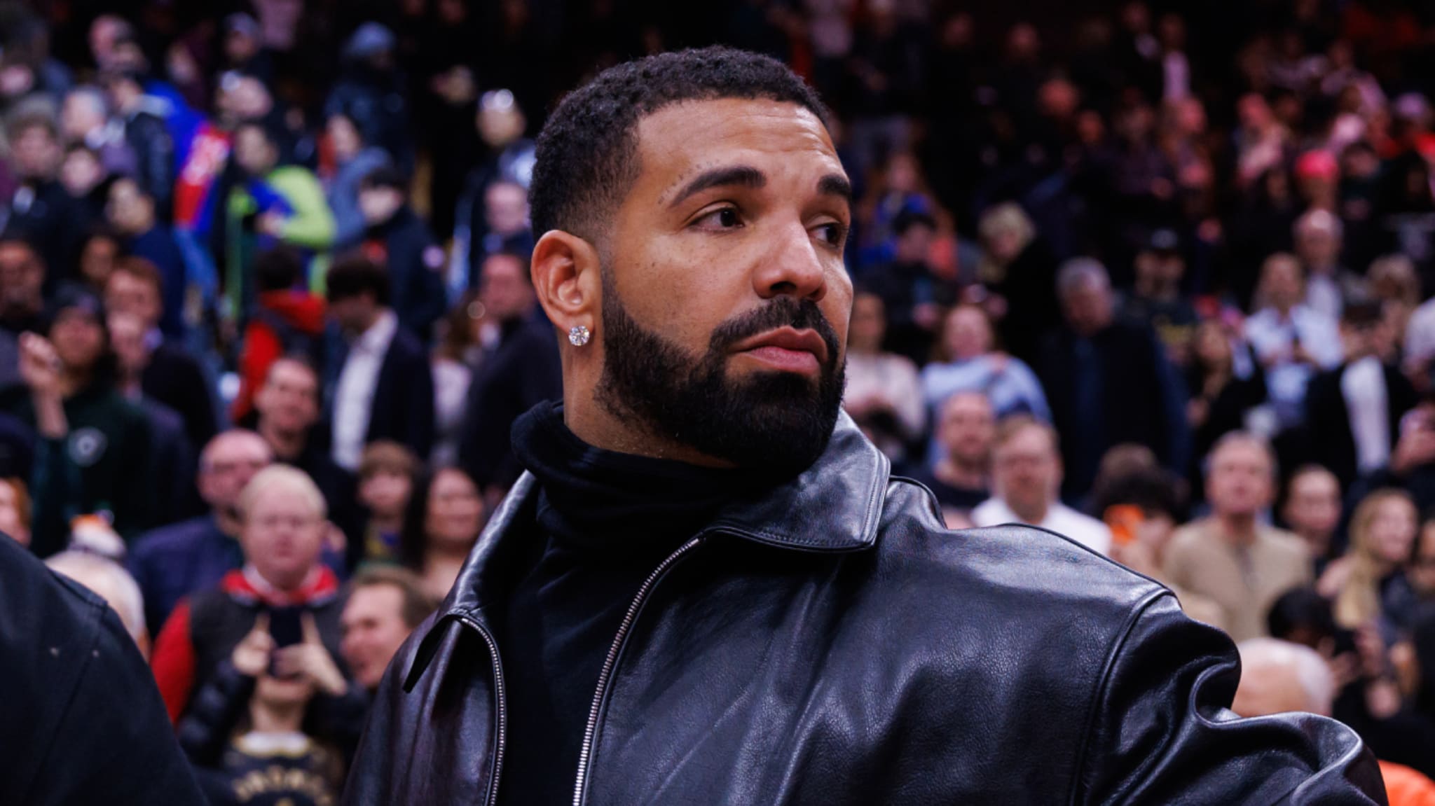 Drake wearing a black leather jacket, standing in a crowded event, looking to the side.