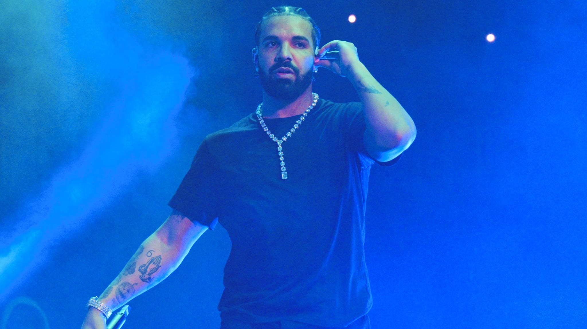 Drake performing on stage, wearing a black shirt and a diamond necklace, with a blue-lit background.