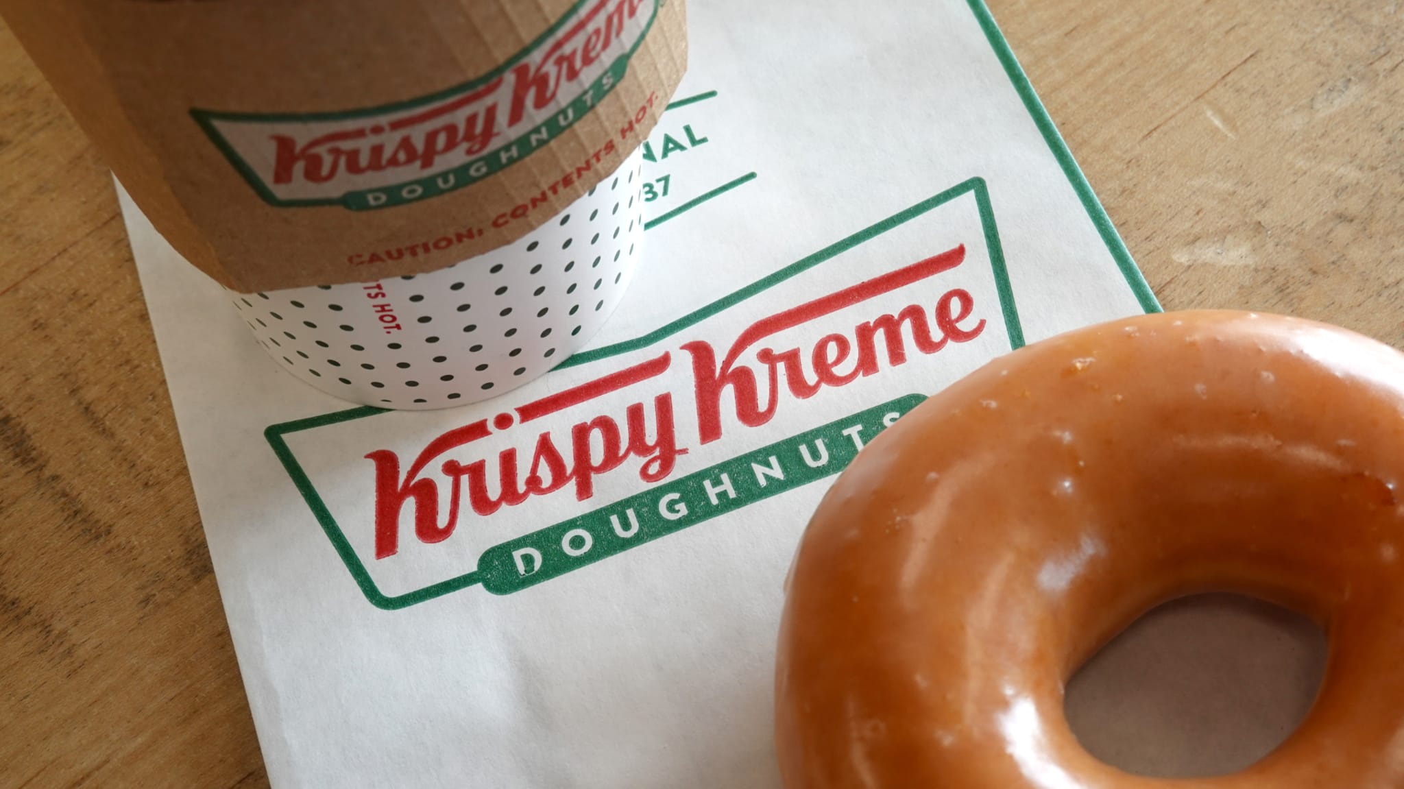 Krispy Kreme coffee cup and glazed doughnut on a branded paper bag on a wooden surface.