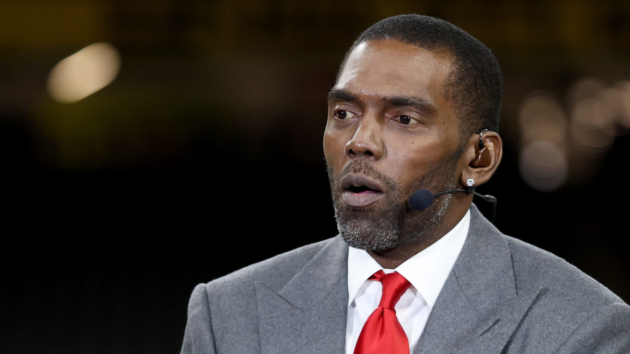 Randy Moss in a gray suit and red tie speaking, wearing a headset microphone.