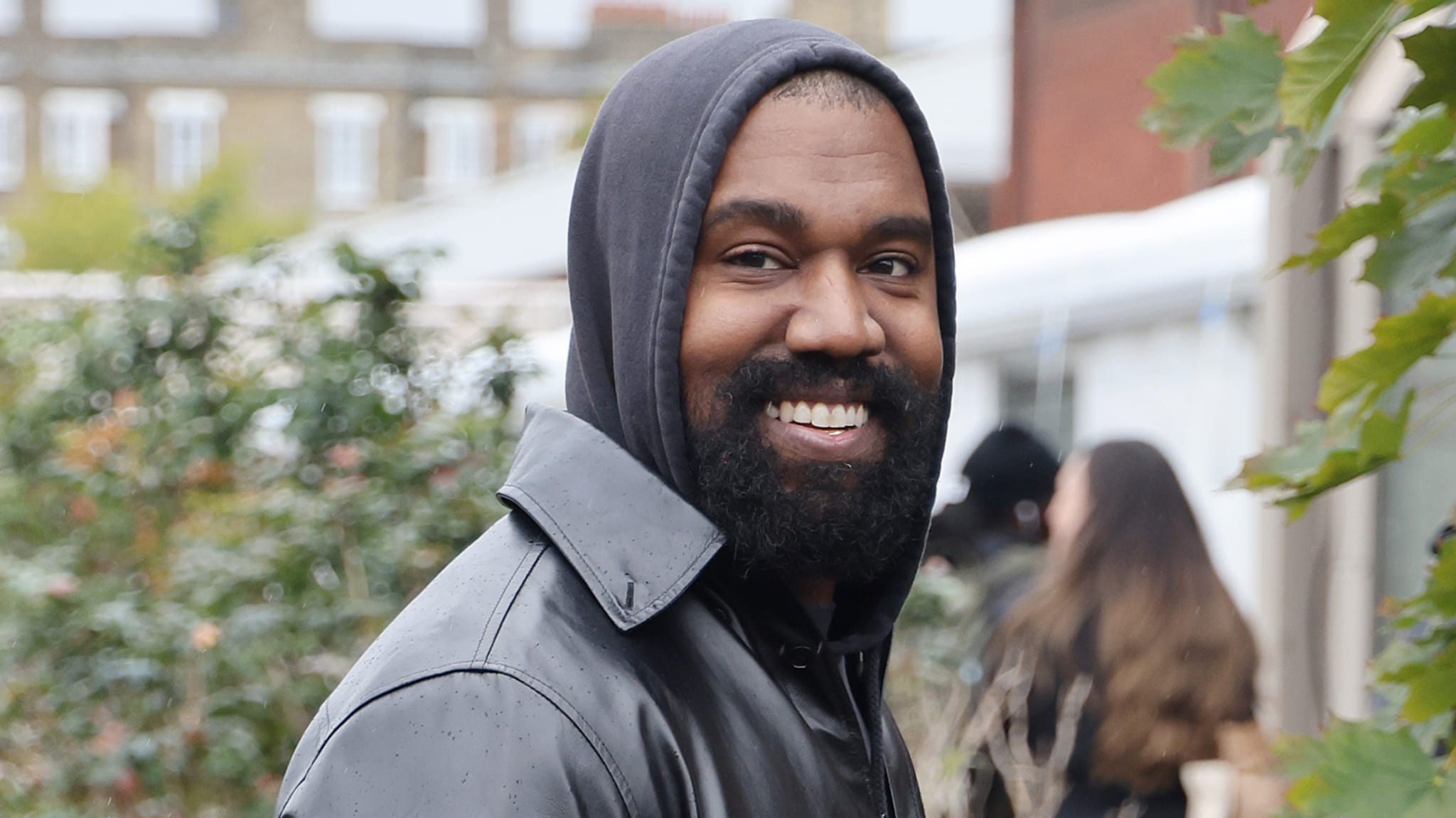 Kanye West smiling, wearing a dark hooded jacket, outdoors with blurred background of greenery and buildings.