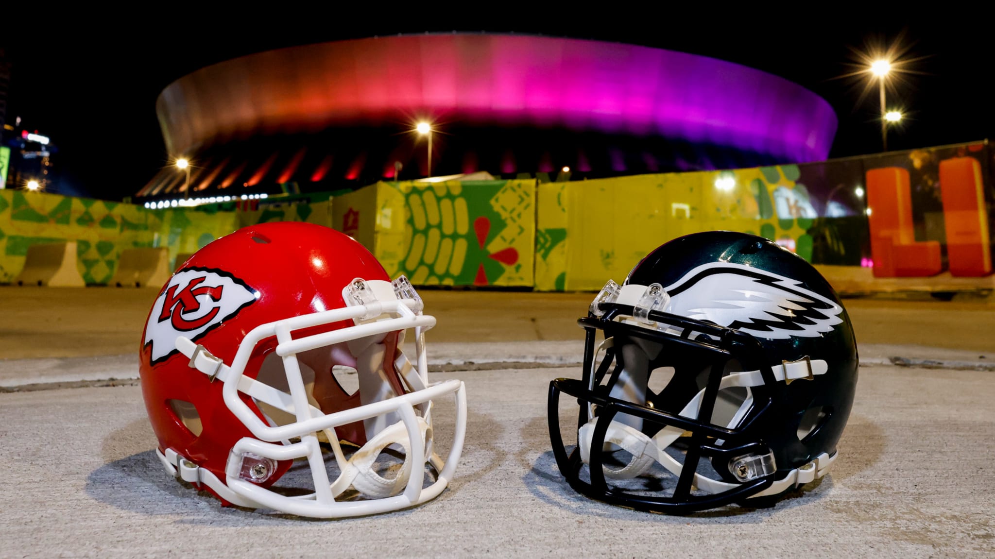 Kansas City Chiefs and Philadelphia Eagles helmets on the ground, with a colorful stadium lit in the background at night.