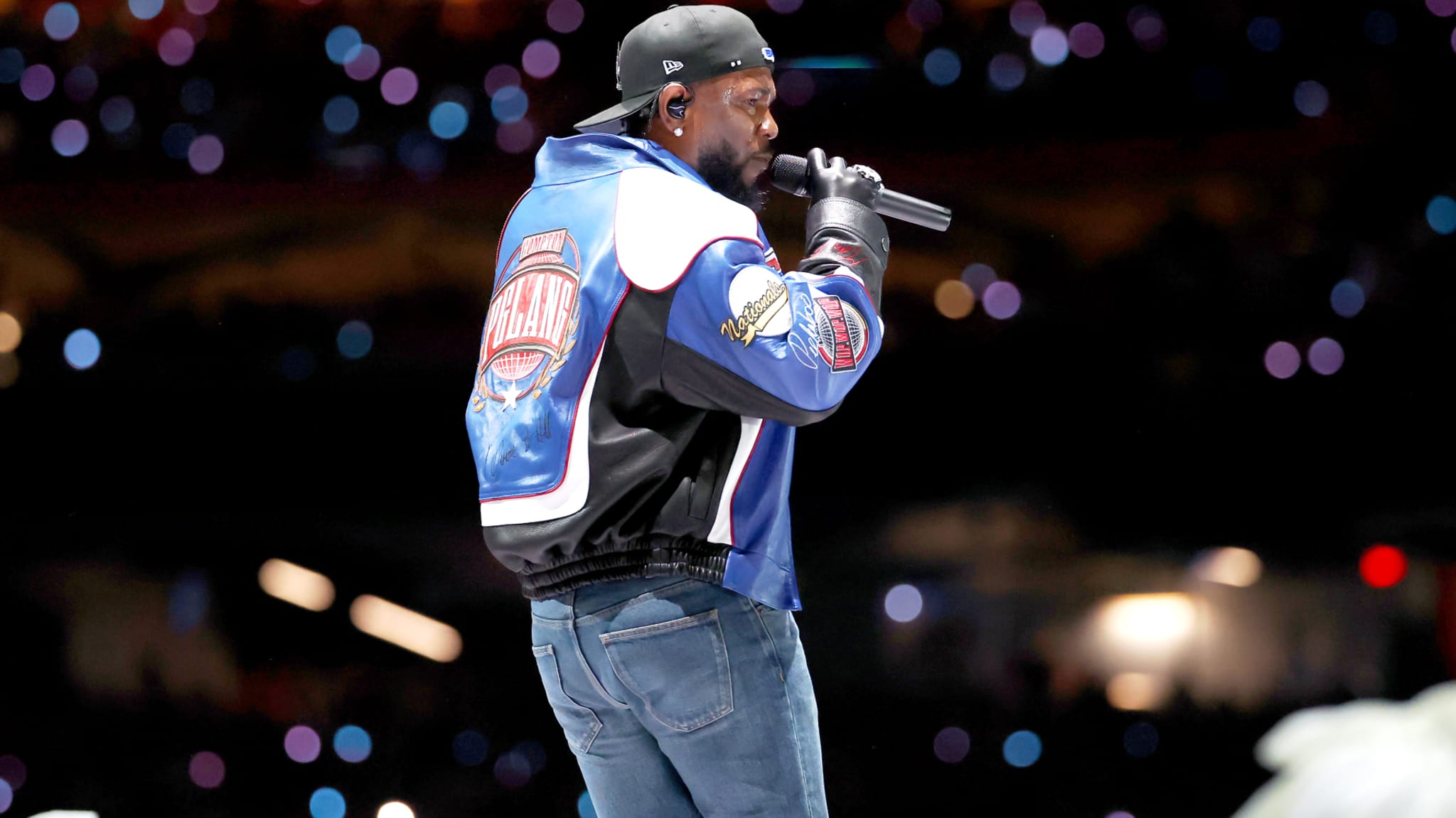A performer on stage wearing a colorful jacket, jeans, and a cap, holding a microphone. The background is filled with blurred lights.