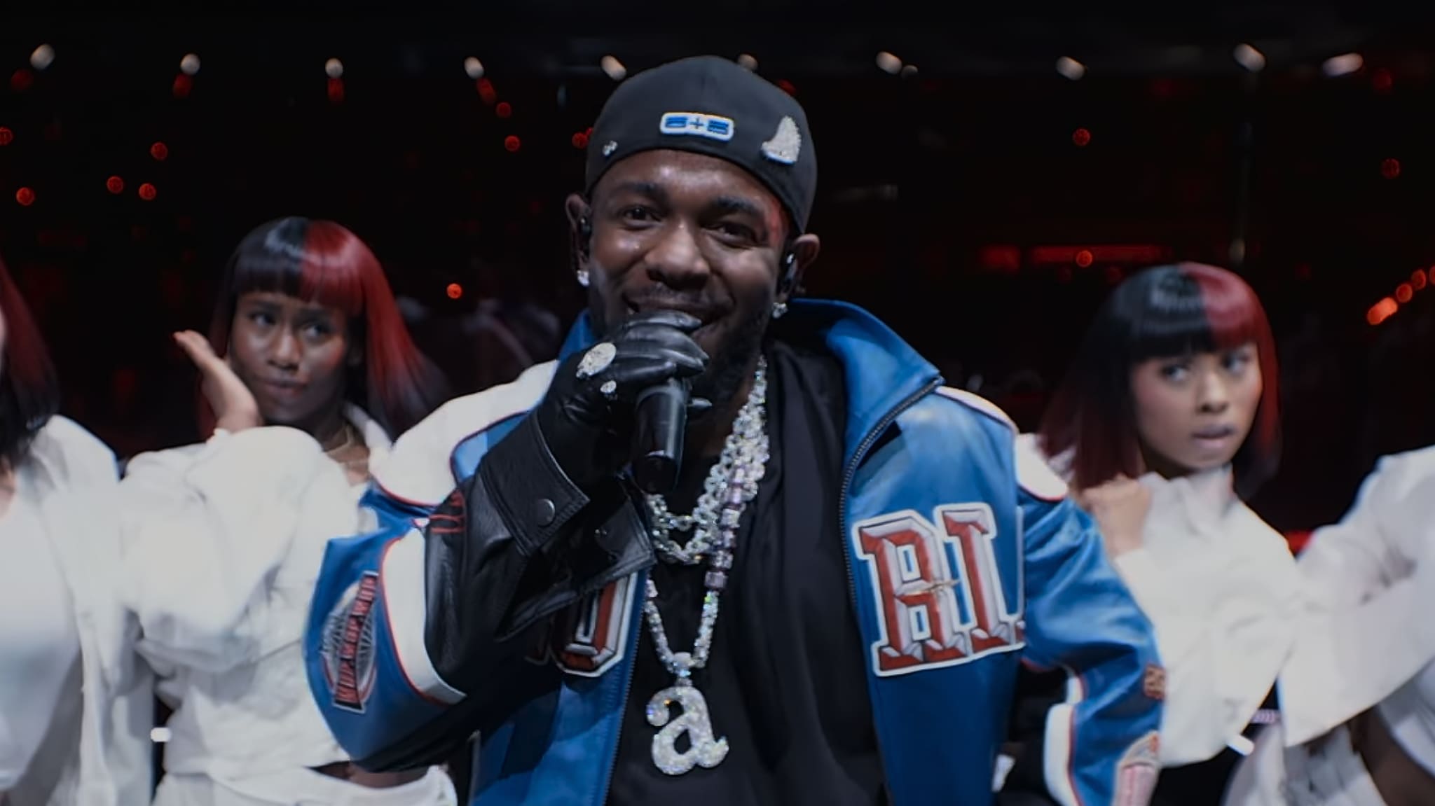 Kendrick Lamar in a blue jacket and cap sings with backup dancers in white outfits on stage.