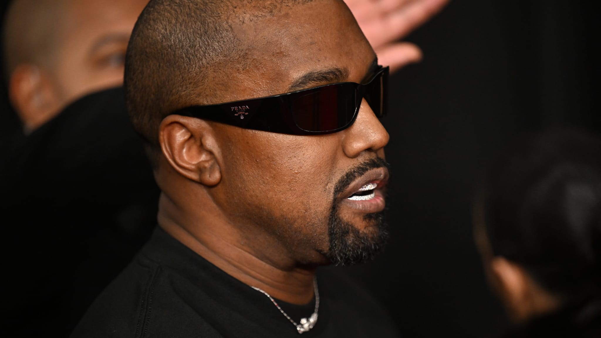 Kanye West wearing black sunglasses and a black shirt, in a side profile view.