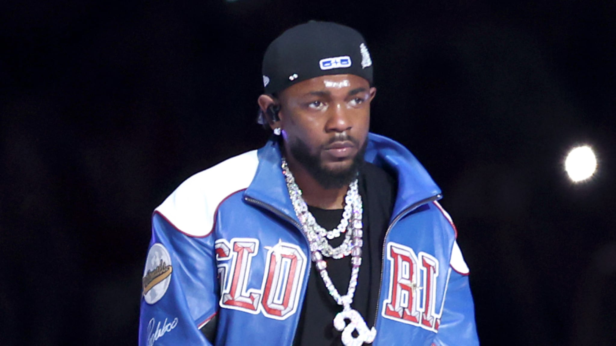 Kendrick Lamar wearing a blue jacket with "Glorious" lettering, a black cap, and multiple necklaces, performing at the Super Bowl LIX Halftime Show.