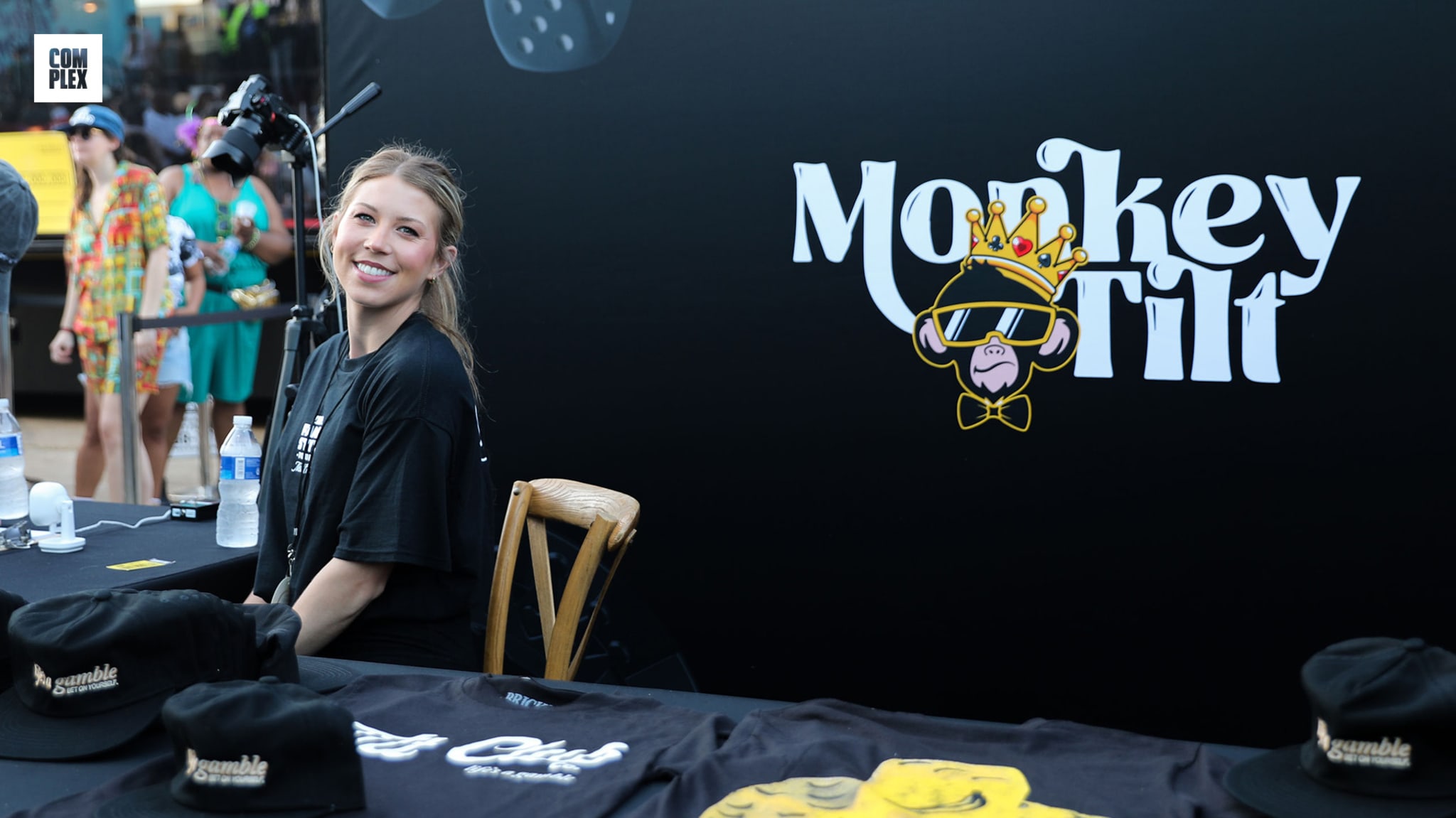 A woman smiling at a booth with "Monkey Tilt" branding, featuring a cartoon monkey with a crown. Merchandise displayed in the foreground.