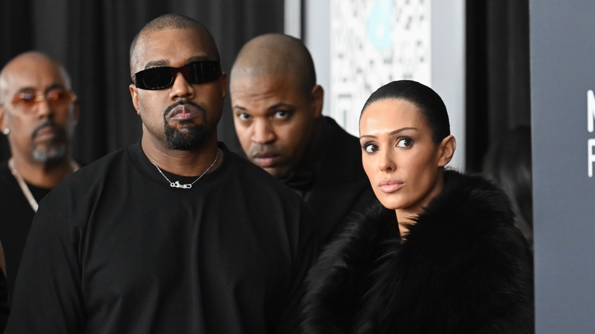 Kanye West in sunglasses and black attire stands beside a woman in a fur coat at an event, with two men in the background.
