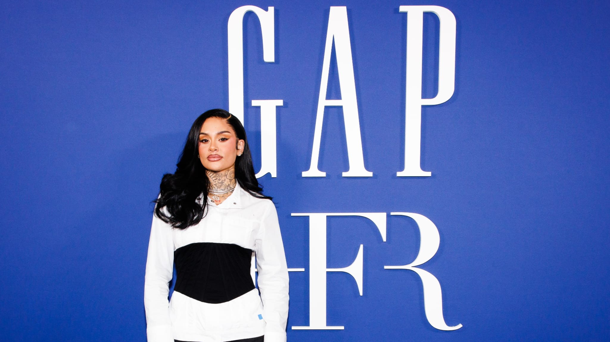 A woman stands in front of a blue background with "GAP FR" text. She wears a white and black outfit with long dark hair.