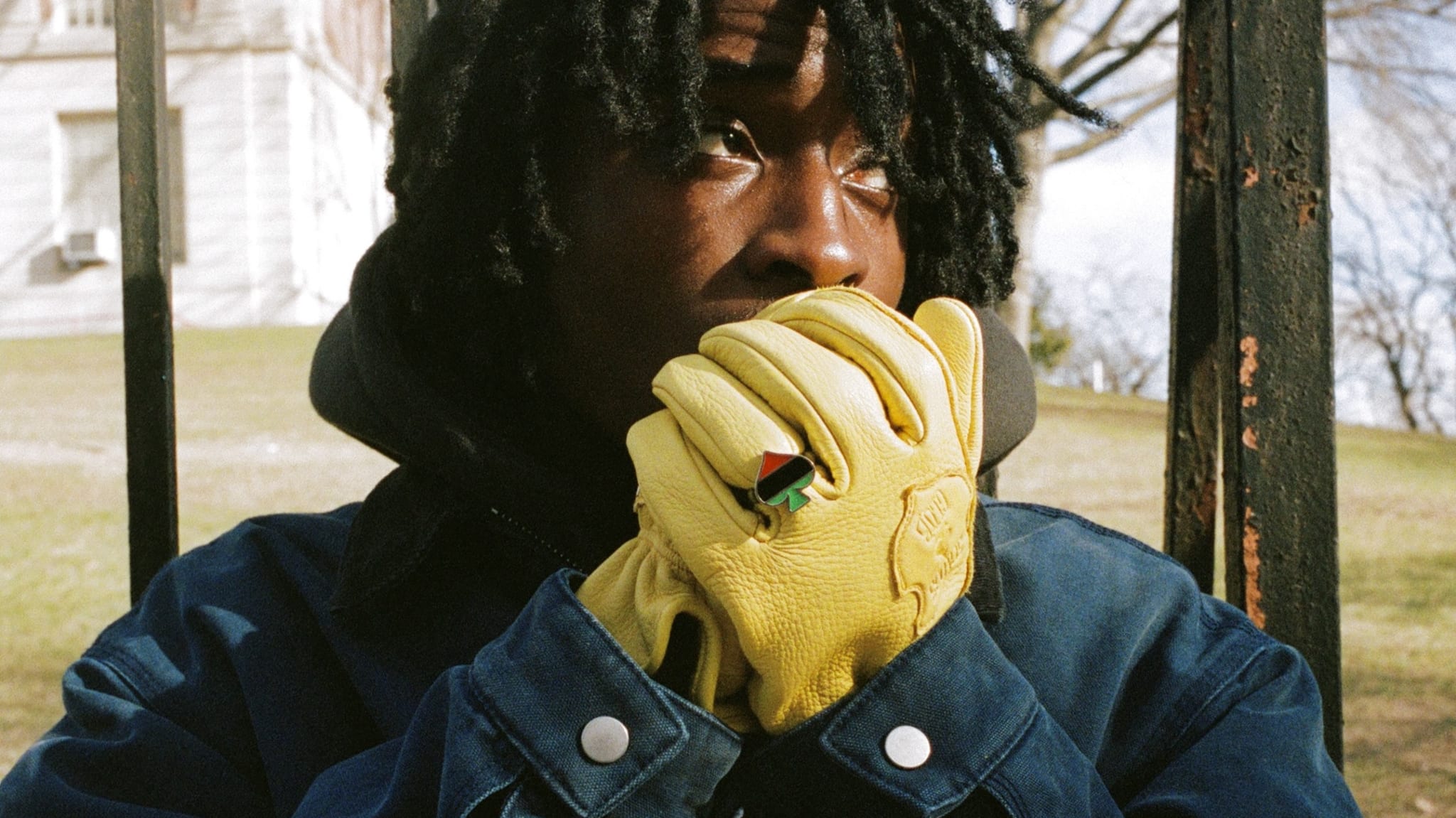 A person with dreadlocks wearing a blue jacket and yellow gloves, looking thoughtfully through a metal fence, with a grassy background.