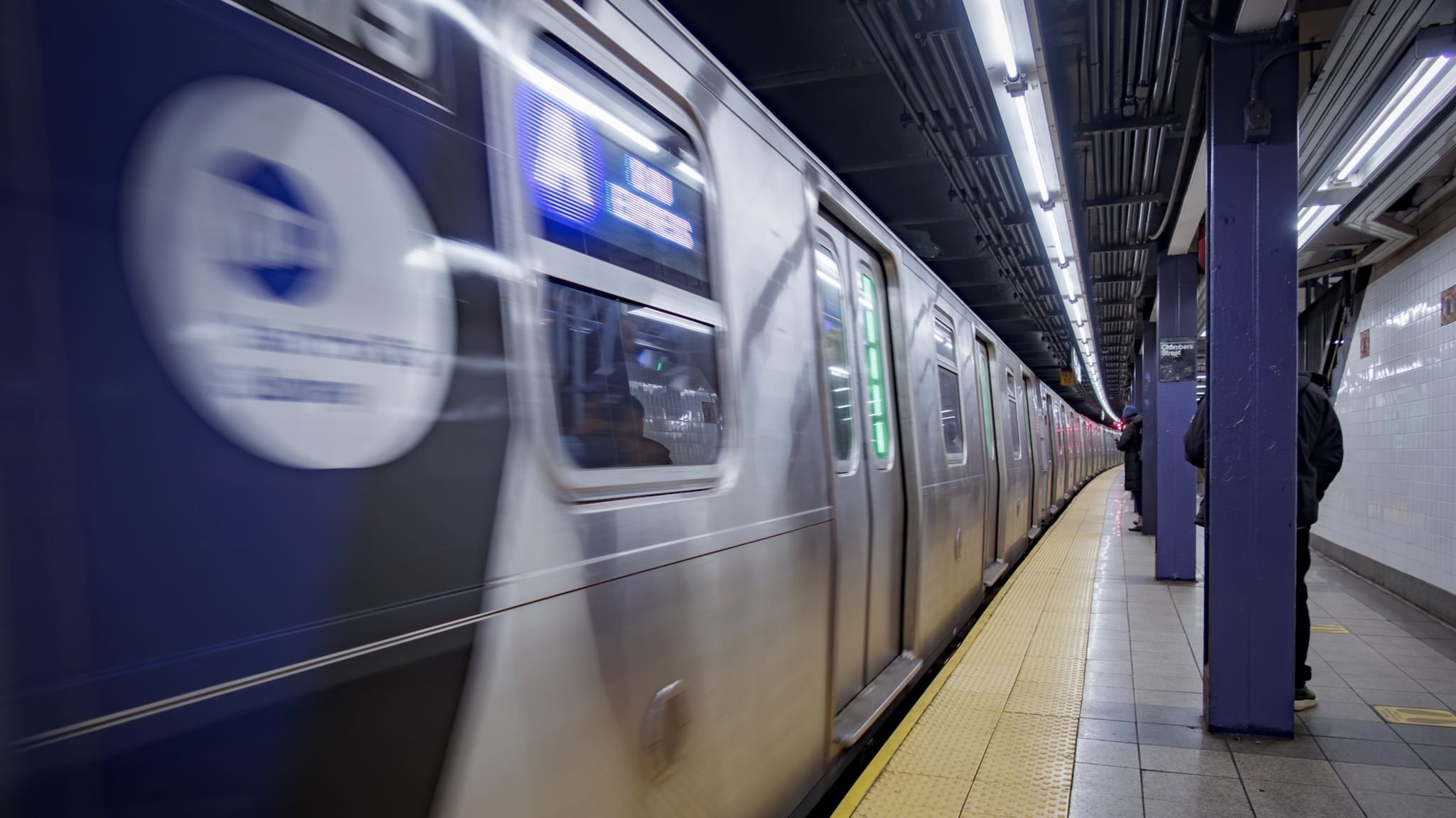 A subway train in motion at a dimly lit underground station, with a person standing on the platform.