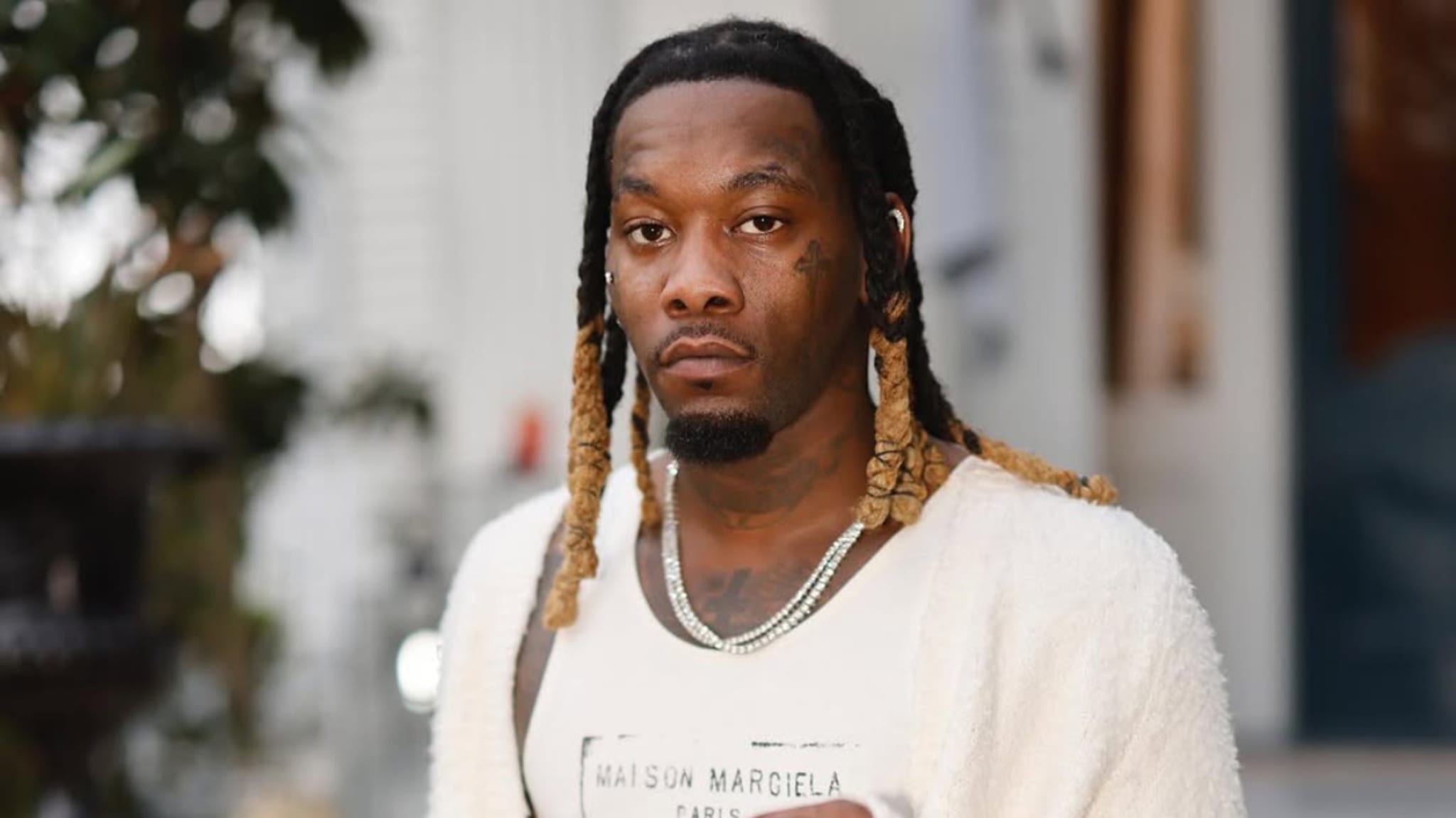 Offset, wearing a Maison Margiela shirt and a white jacket, with braided hair and jewelry, standing outdoors.