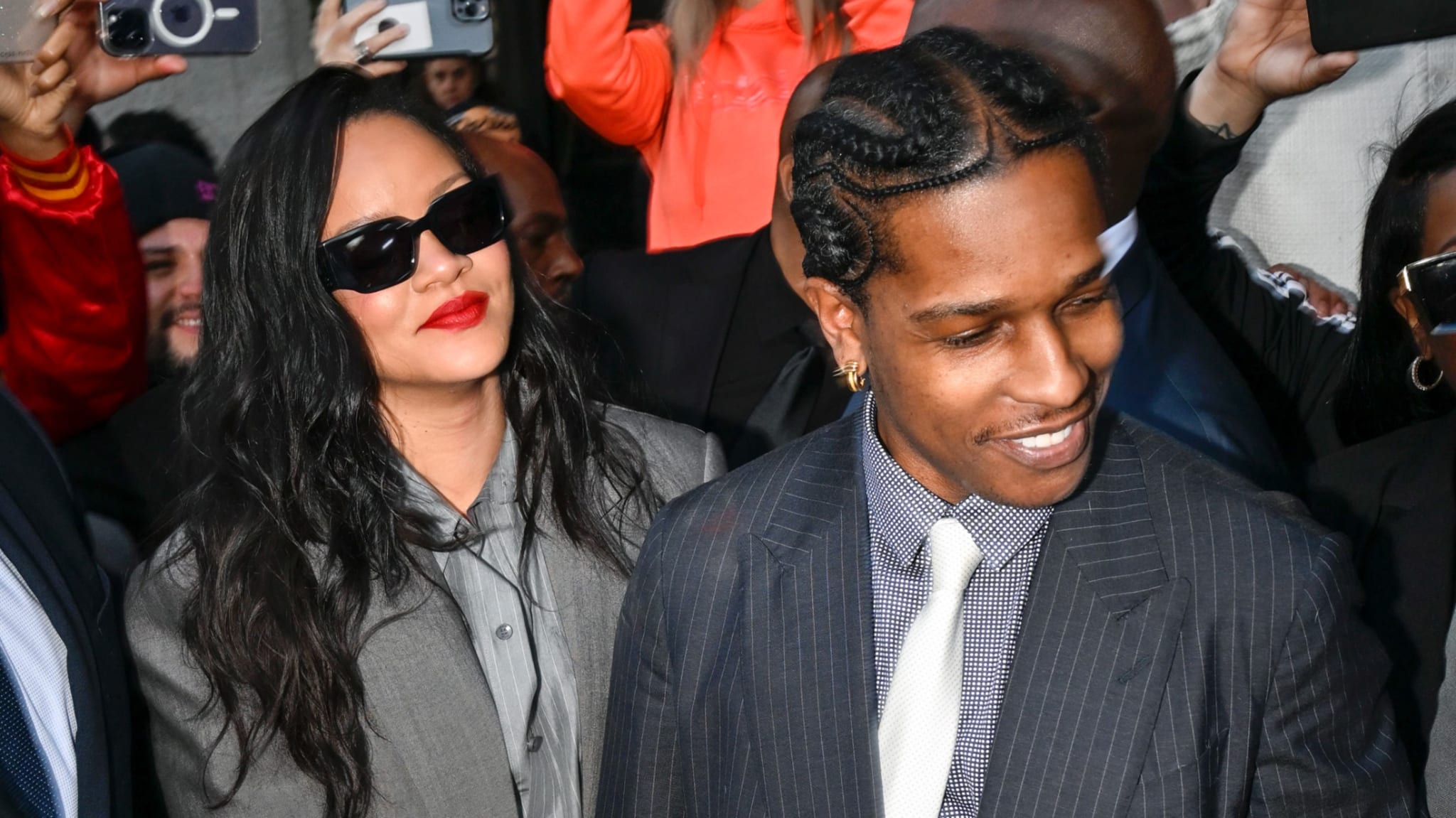 Rihanna and A$AP Rocky in stylish outfits, surrounded by a crowd. Rihanna wears sunglasses and a grey suit; A$AP Rocky is in a pinstripe suit.