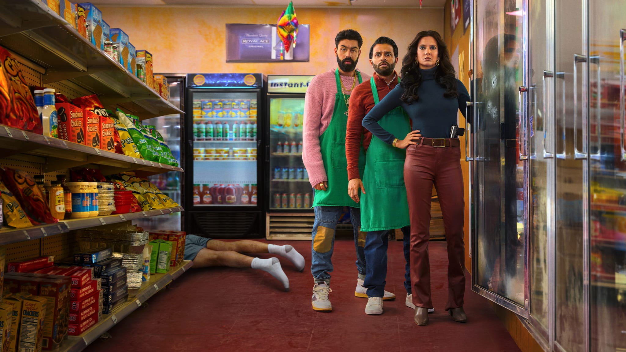 Three people in a convenience store stand in front of a freezer, with a pair of legs sticking out from an aisle. Shelves of snacks are visible.