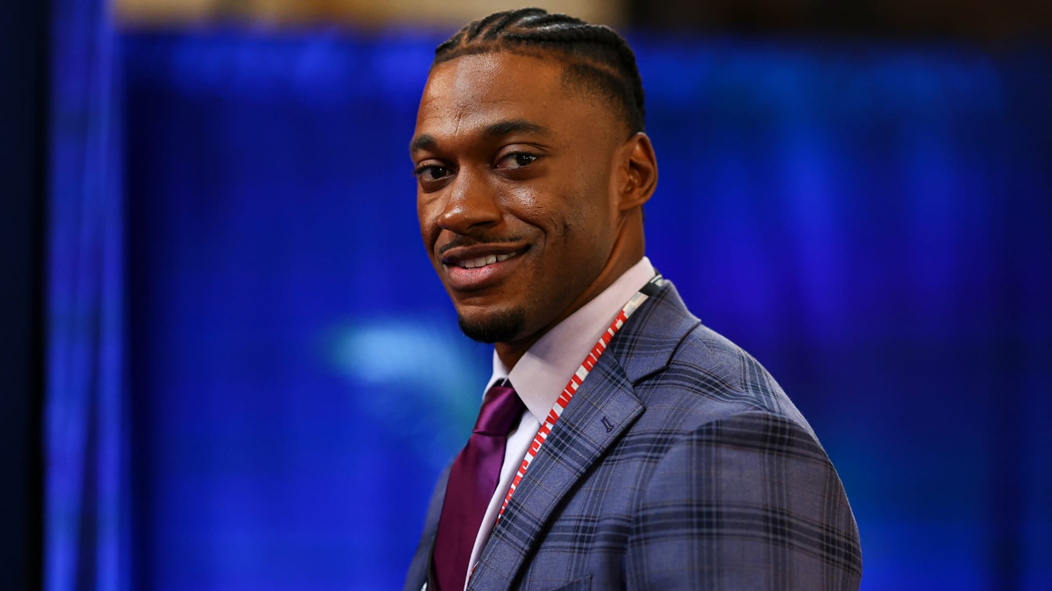 A man in a checkered suit and tie, smiling, with braided hair, against a blurred blue background.