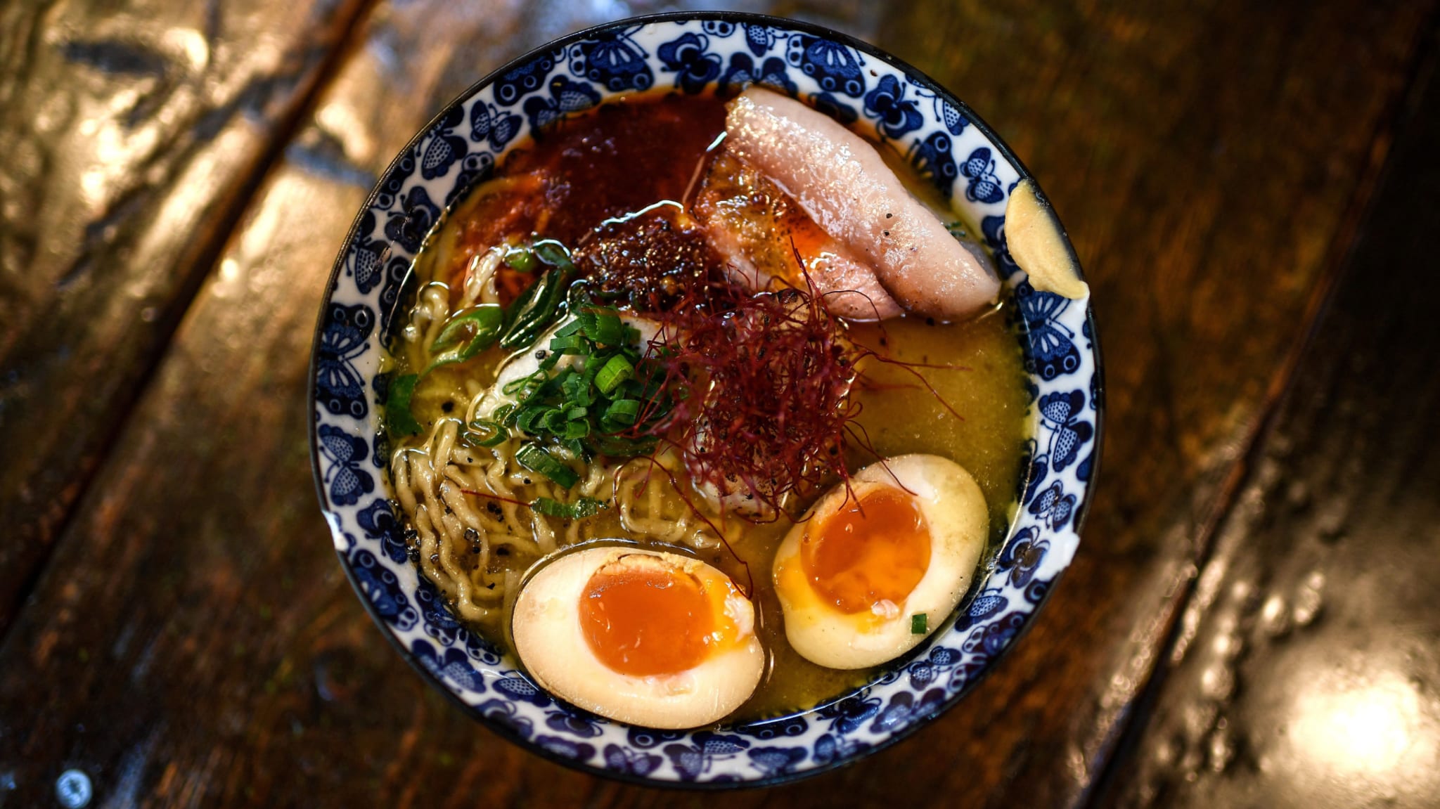 A bowl of ramen with sliced pork, soft-boiled eggs, green onions, and red chili threads in a decorative blue and white bowl.