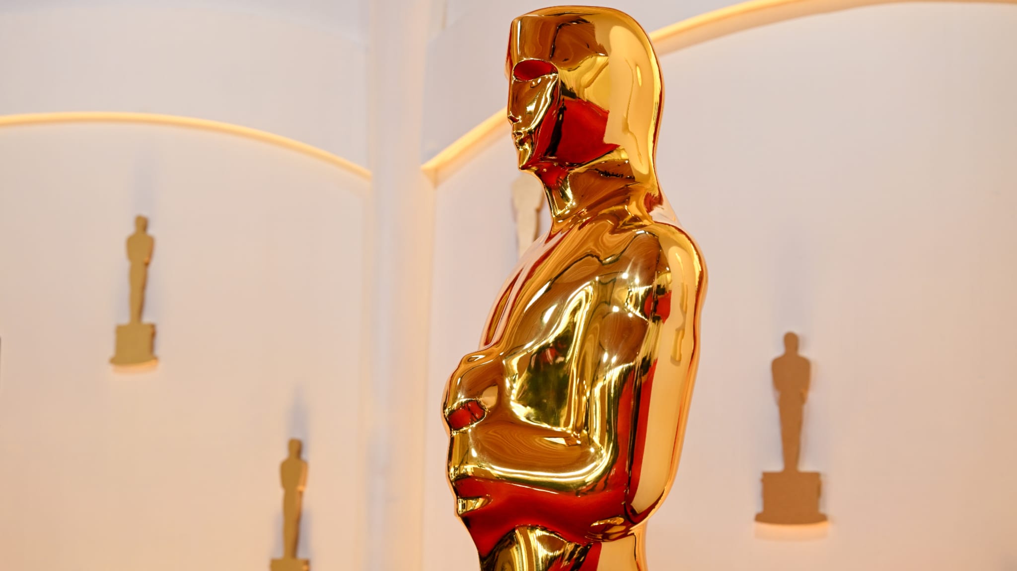 A large golden Oscar statue on display, with smaller Oscar statues in the background, set against a white wall.