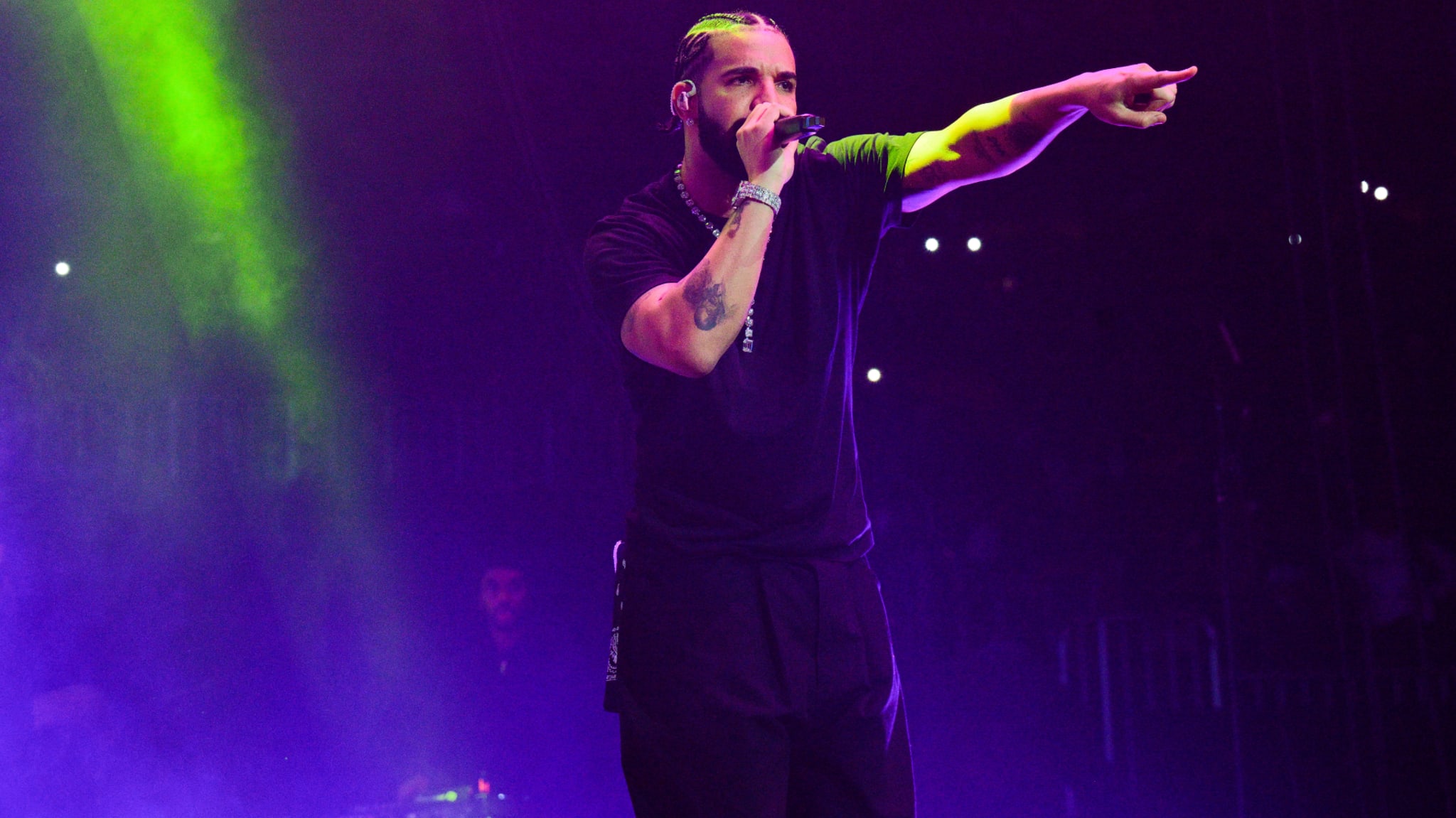 Drake performing on stage, wearing a black outfit, with vibrant purple and green lighting in the background.