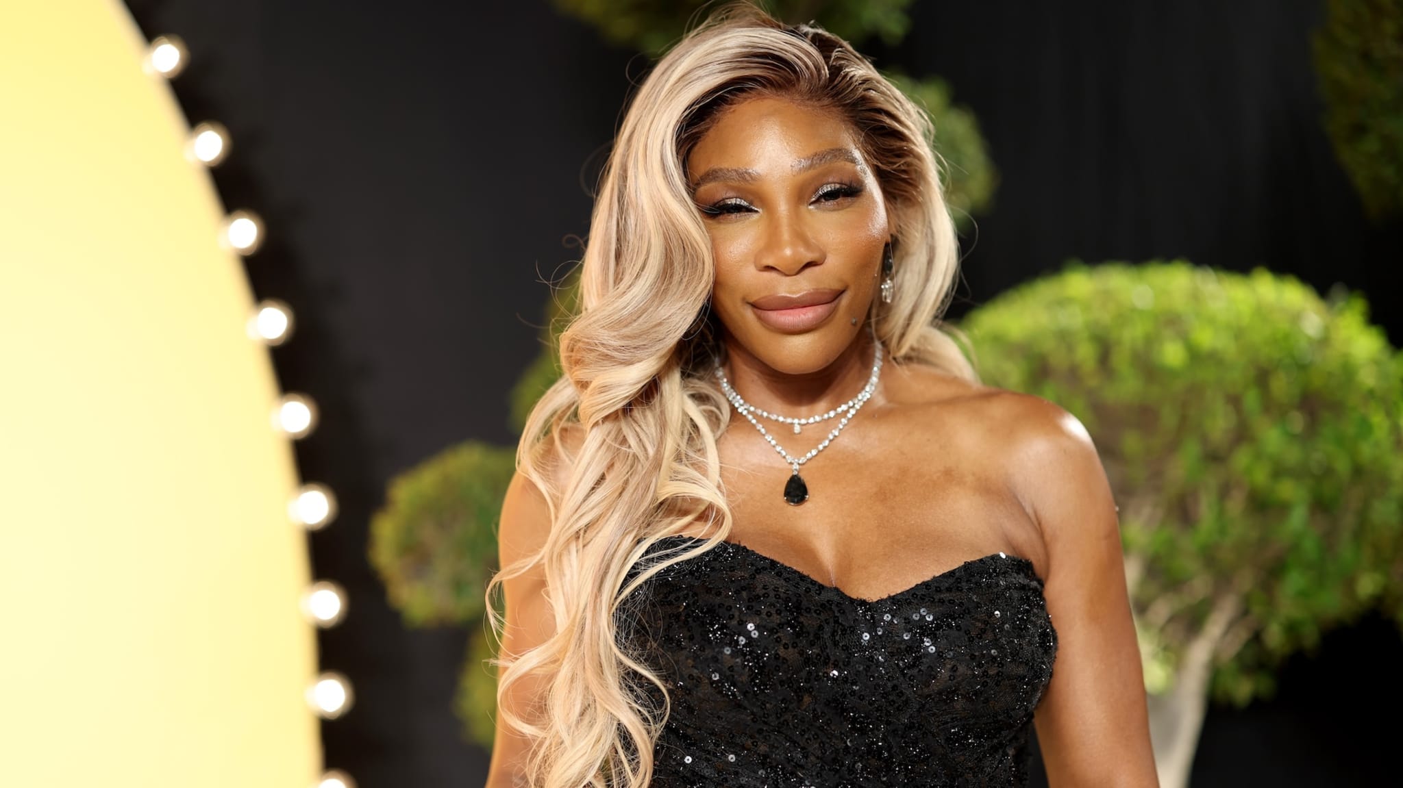 Serena Williams in a black sequined dress, with long blonde hair and a necklace, standing in front of a blurred background.